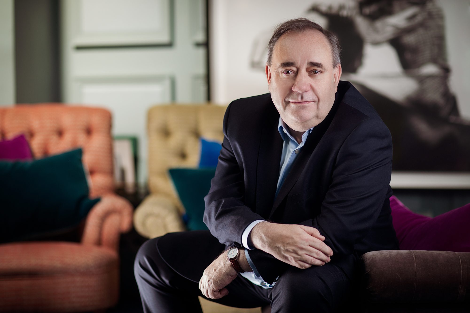 Alex Salmond seated on the arm of a sofa, in a dark suit and open-necked shirt