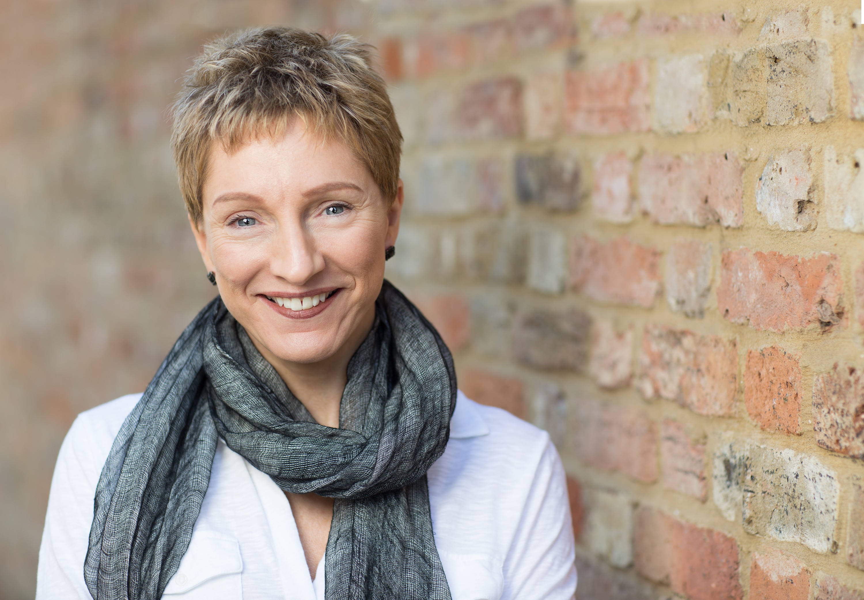Isobel McDermott-Spencer with short fair hair, in a white shirt and grey scarf, smiling beside an old brick wall, portrait by Mat Smith Photography