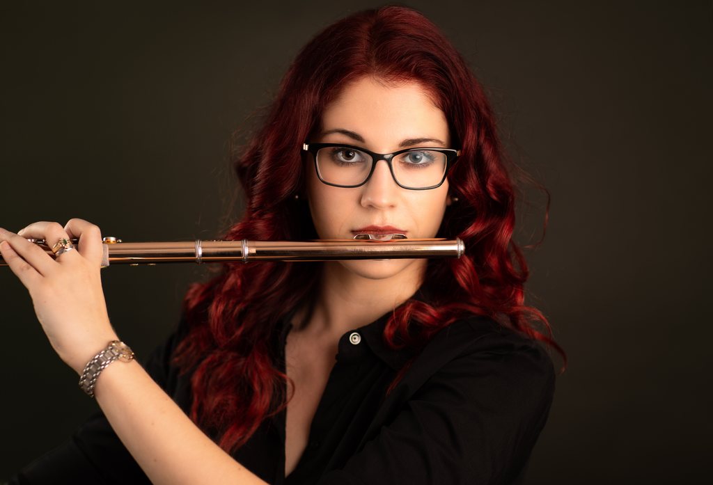 Lindsay Bryden, flautist, with long red hair and glasses, holding her flute to her lips against a dark background, portrait by Mat Smith Photography, London