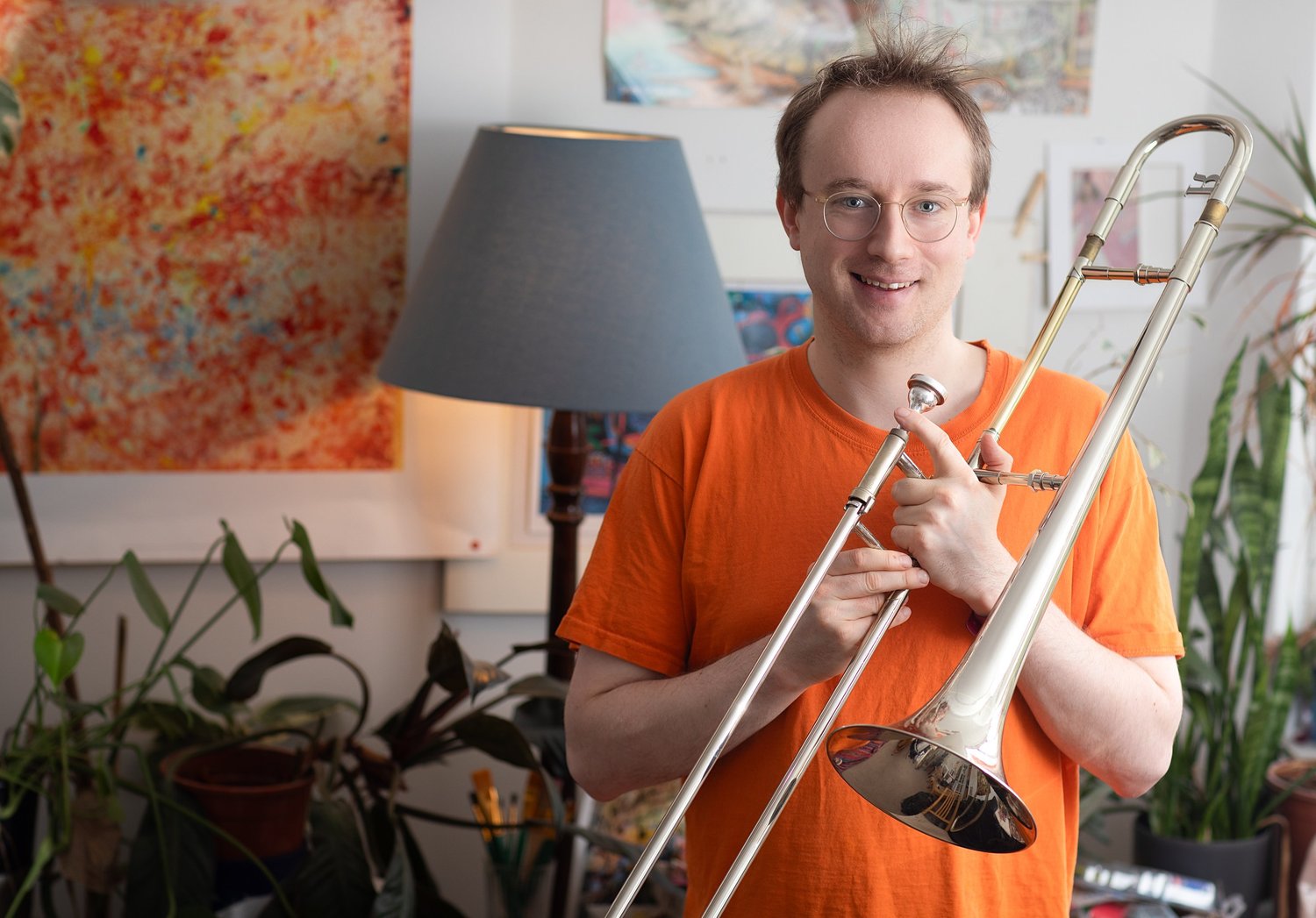 Alex Paxton, composer and trombonist, holding his trombone at home among plants and paintings, portrait by Mat Smith Photography