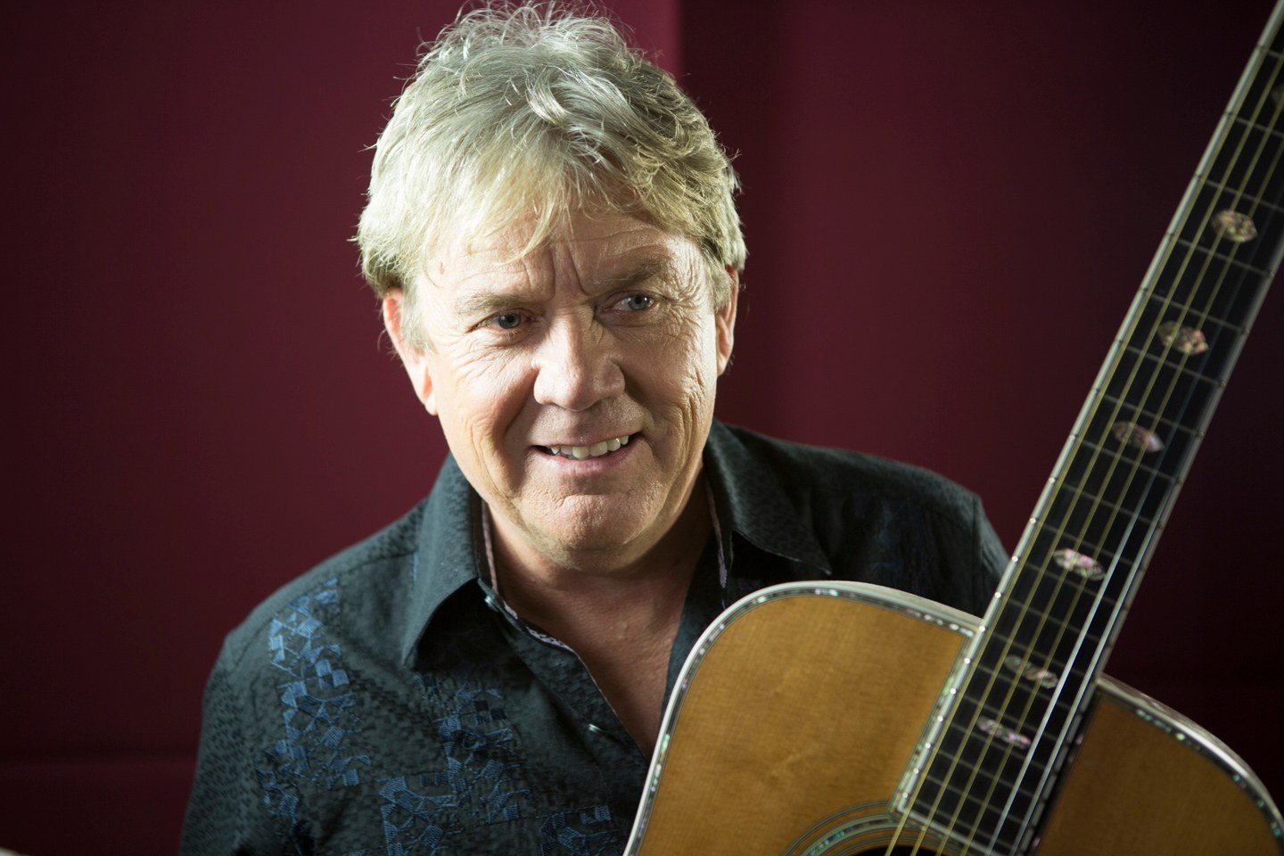 Mike Stock, songwriter and producer, with an acoustic guitar against a deep red wall, portrait by Mat Smith Photography