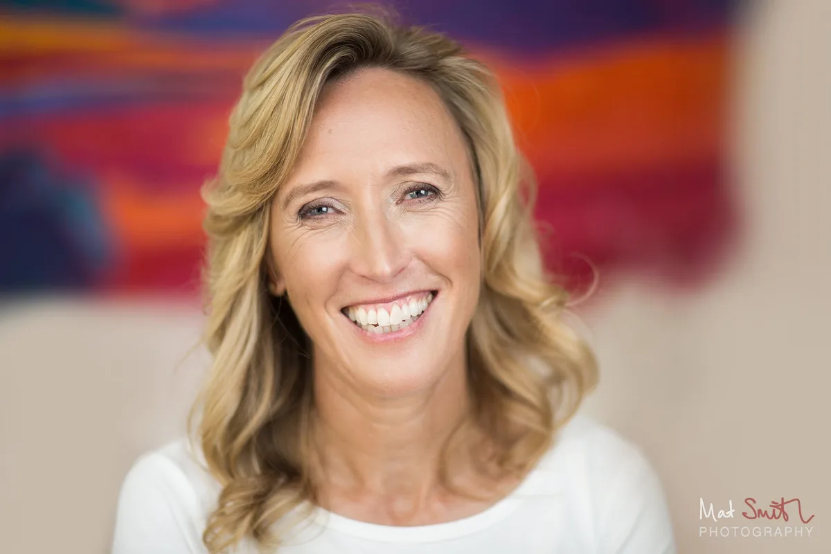 Smiling woman with long wavy blonde hair in a white top in front of a brightly coloured painting, portrait by Mat Smith Photography, London.