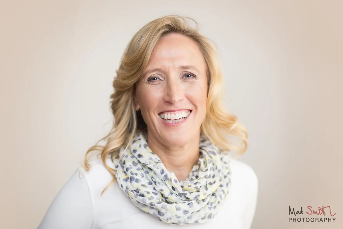 Smiling woman with long wavy blonde hair in a white top and spotted scarf against a plain cream wall, portrait by Mat Smith Photography, London.