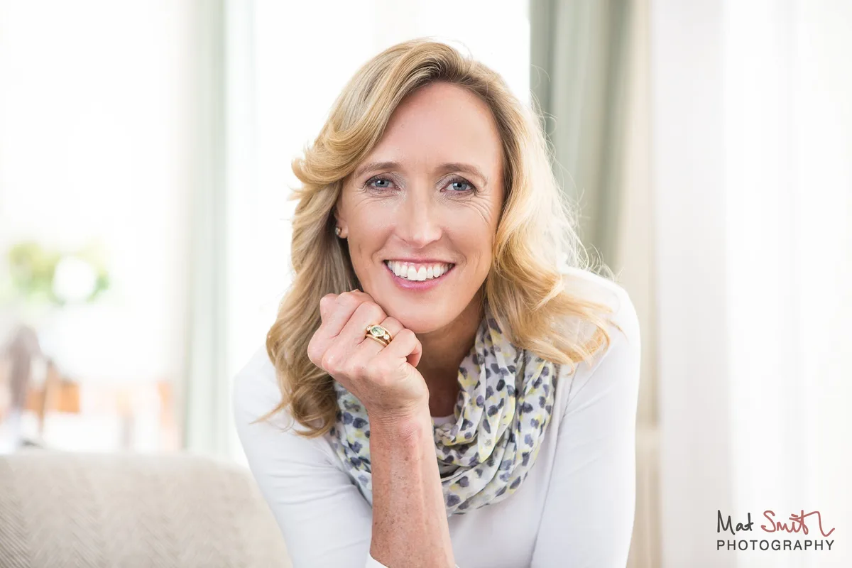 Woman with long wavy blonde hair resting her chin on her hand by a bright window, in a white top and spotted scarf, portrait by Mat Smith Photography, London.