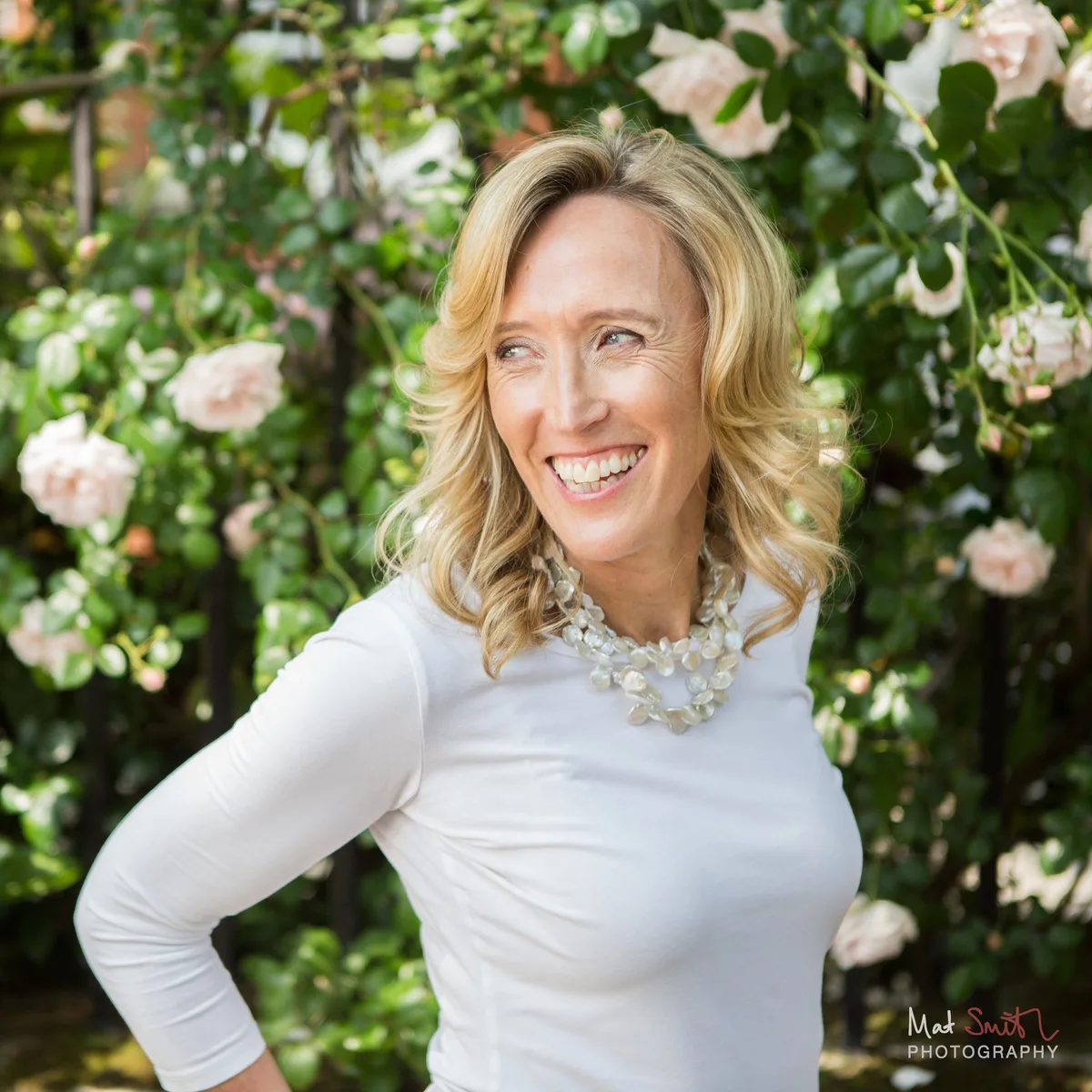 Woman laughing and looking to the side with one hand on her hip, in front of pale pink climbing roses, portrait by Mat Smith Photography, London.