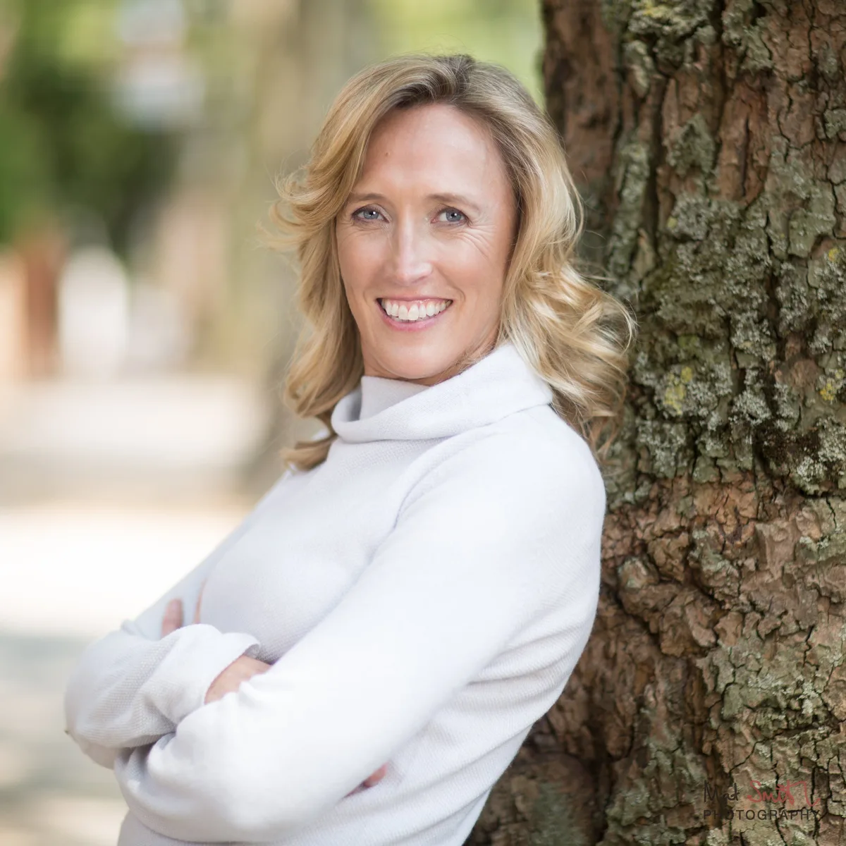 Smiling woman in a white roll neck jumper leaning against a large tree trunk with her arms folded, portrait by Mat Smith Photography, London.