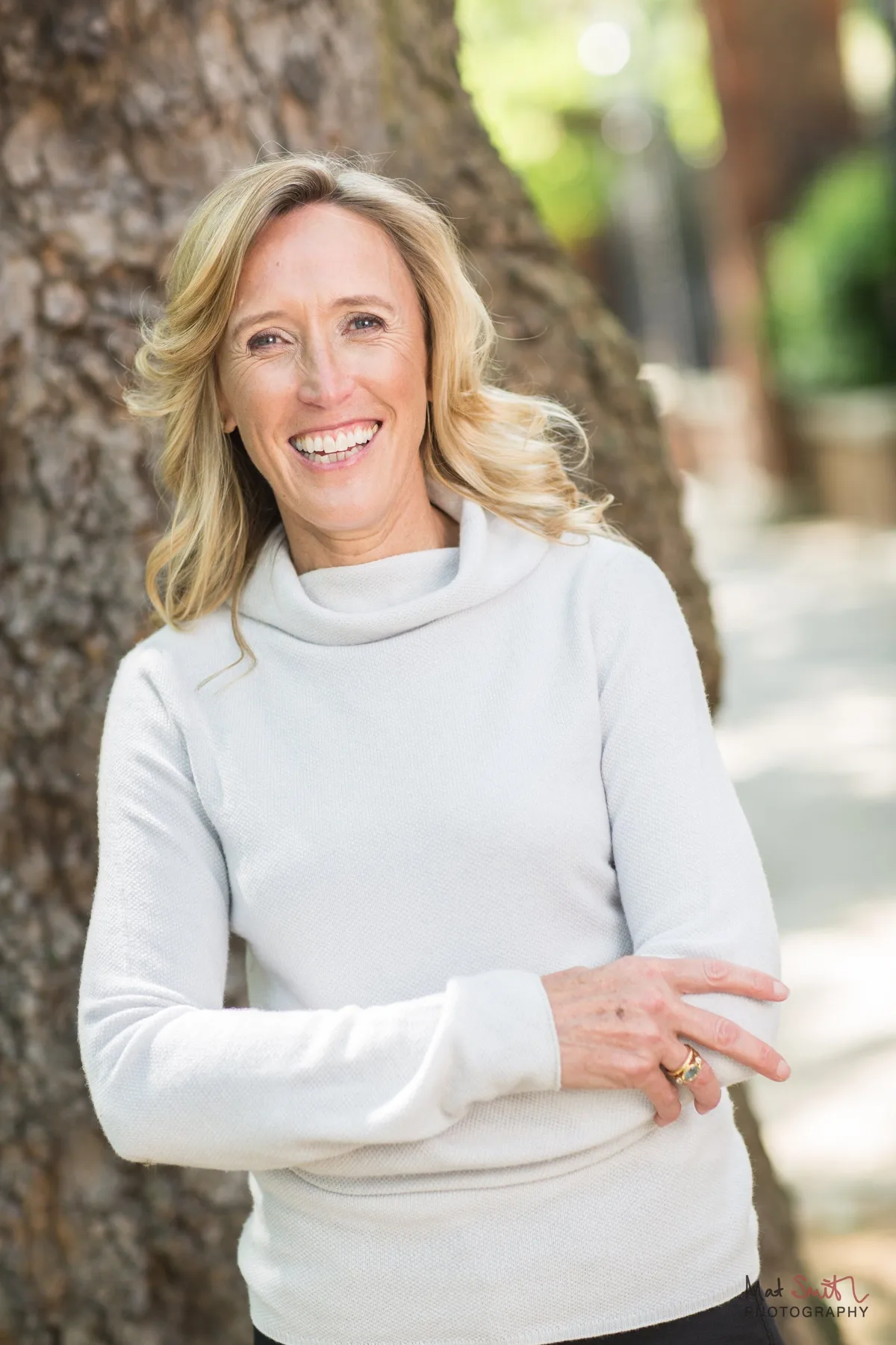 Upright portrait of a smiling woman in a white roll neck jumper leaning against a tree on a leafy pavement, portrait by Mat Smith Photography, London.