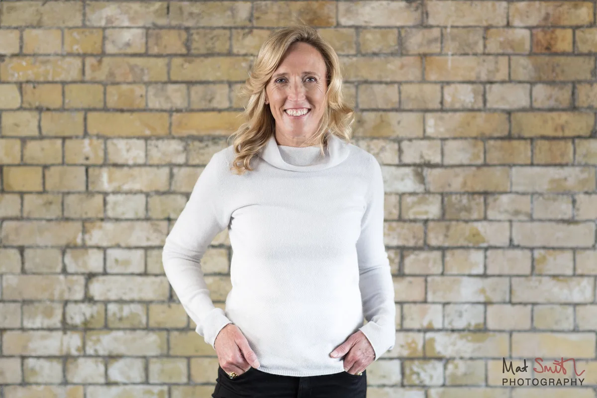Smiling woman in a white jumper and black trousers standing with her thumbs in her pockets against a yellow brick wall, portrait by Mat Smith Photography, London.