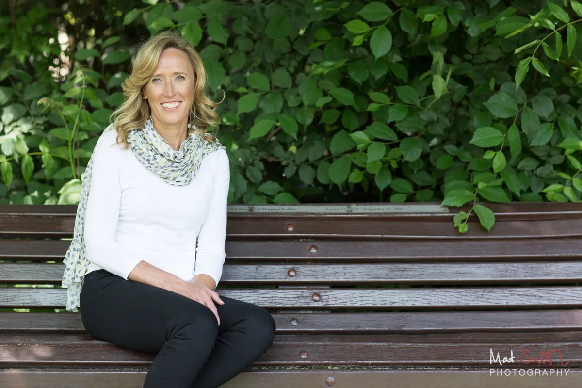 Smiling woman in a white top and scarf sitting on a wooden bench in front of green foliage, portrait by Mat Smith Photography, London.
