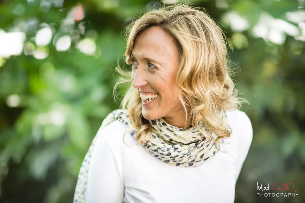 Close portrait of a woman laughing and looking away, in a white top and patterned scarf with green leaves behind, portrait by Mat Smith Photography, London.