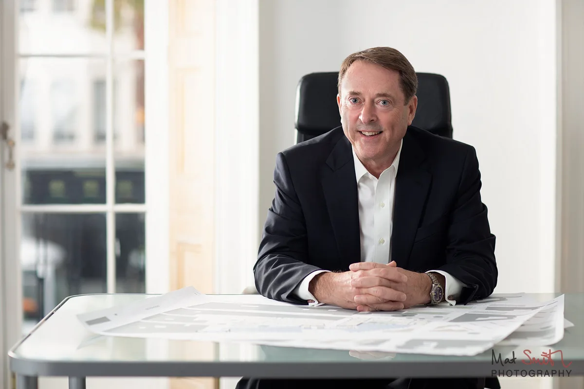 A smiling man in a dark suit and open white shirt sits with hands clasped at a glass desk covered in plans, by a tall window, portrait by Mat Smith Photography, London
