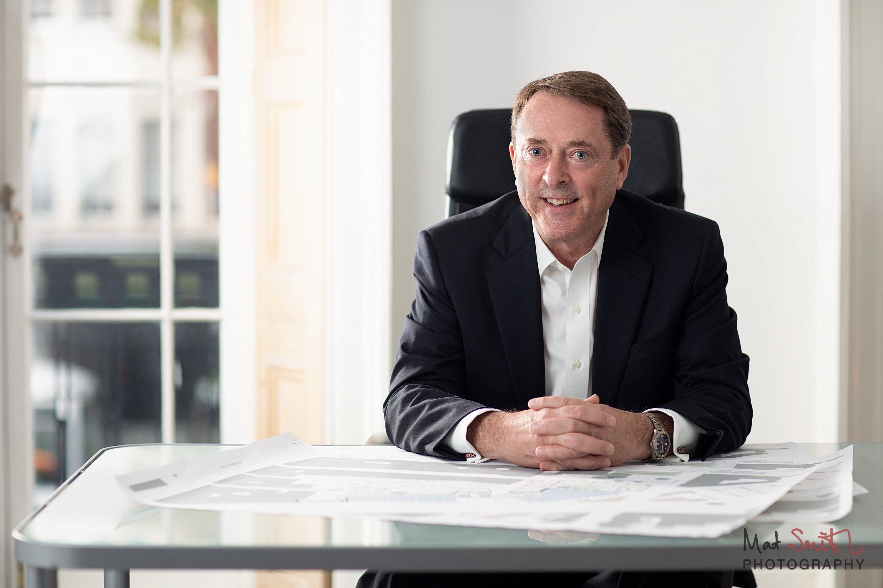 A smiling man in a dark suit and open white shirt sits with hands clasped at a glass desk covered in plans, by a tall window, portrait by Mat Smith Photography, London