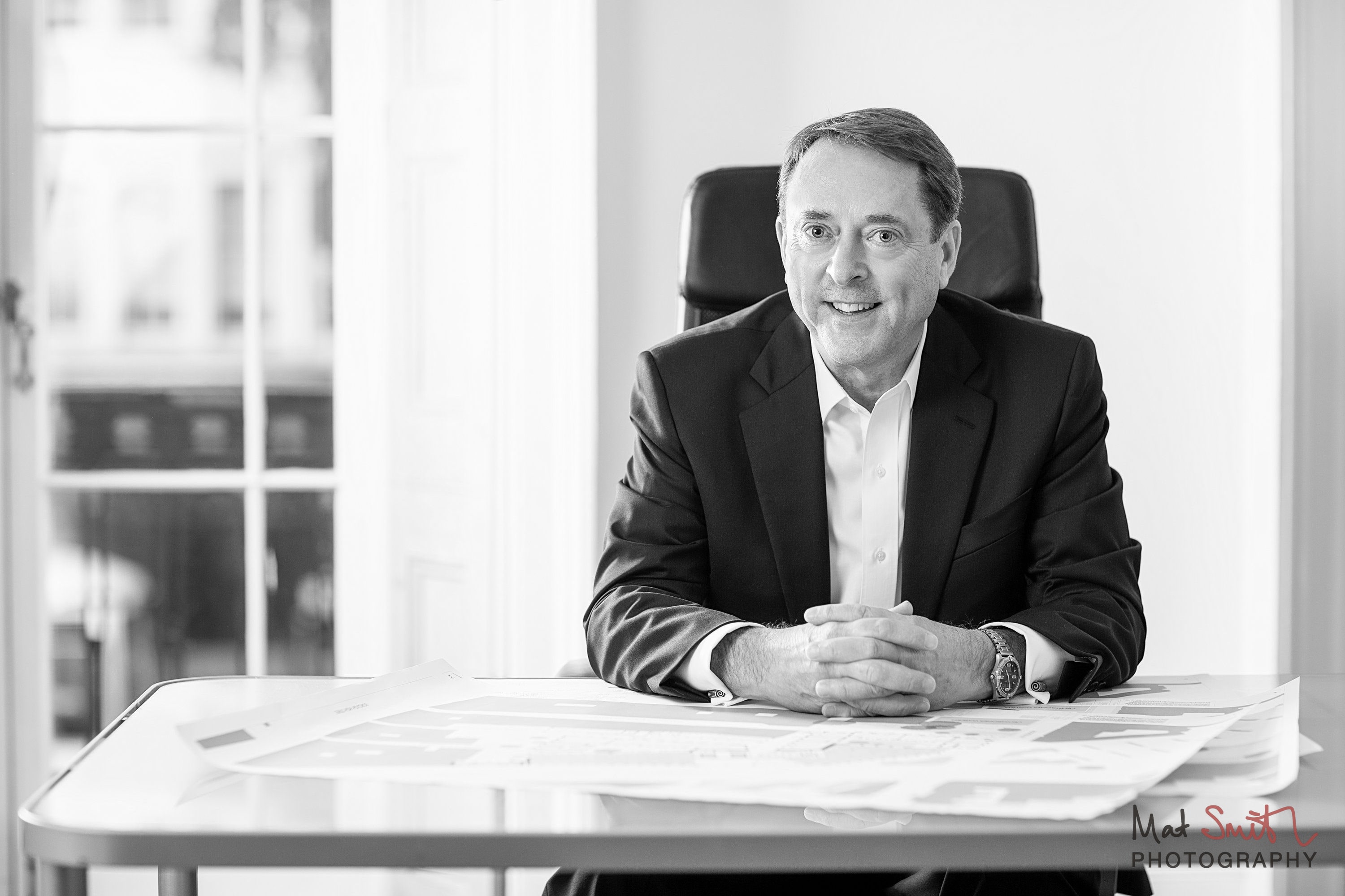 Black and white portrait of a man in a dark suit seated with hands clasped at a desk covered in plans, portrait by Mat Smith Photography, London