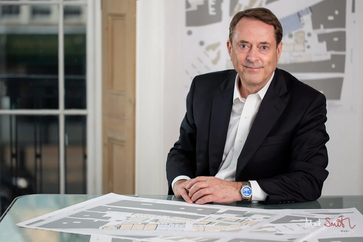 A man in a dark suit leans on a glass desk spread with site plans, with a plan pinned to the wall behind, portrait by Mat Smith Photography, London