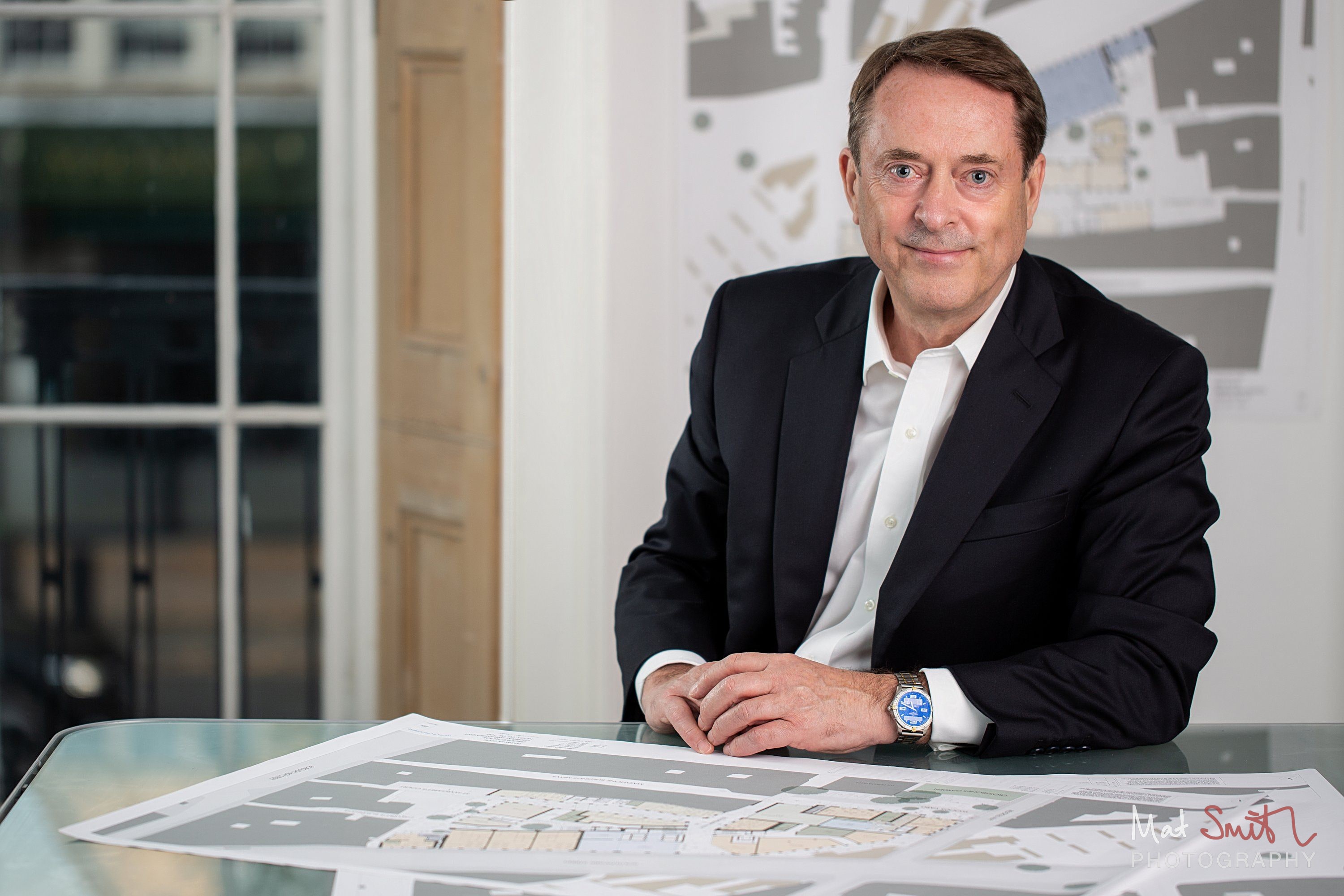 A man in a dark suit leans on a glass desk spread with site plans, with a plan pinned to the wall behind, portrait by Mat Smith Photography, London