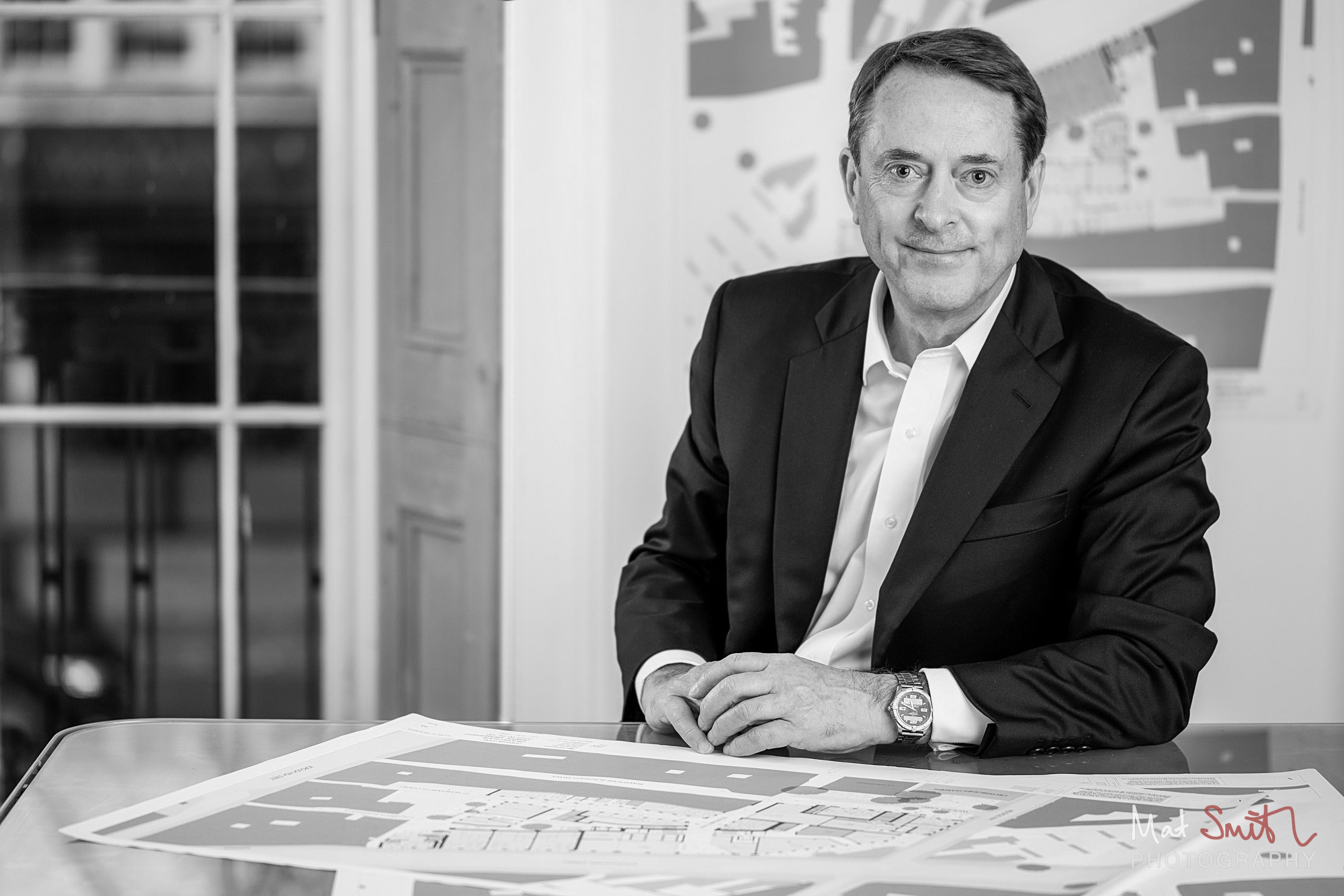 Black and white image of a man in a suit leaning on a desk of site plans, smiling at the camera, portrait by Mat Smith Photography, London