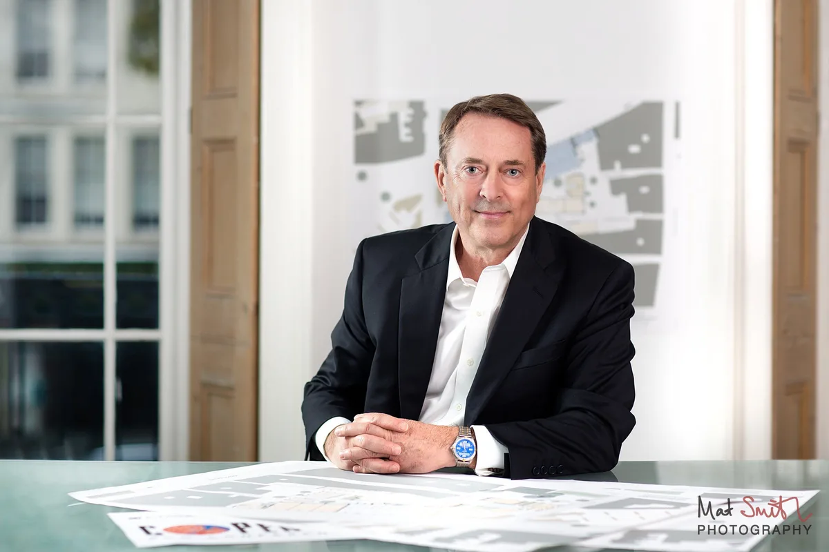 A man in a dark suit sits at a glass desk with printed plans in a bright office with tall windows and wooden shutters, portrait by Mat Smith Photography, London