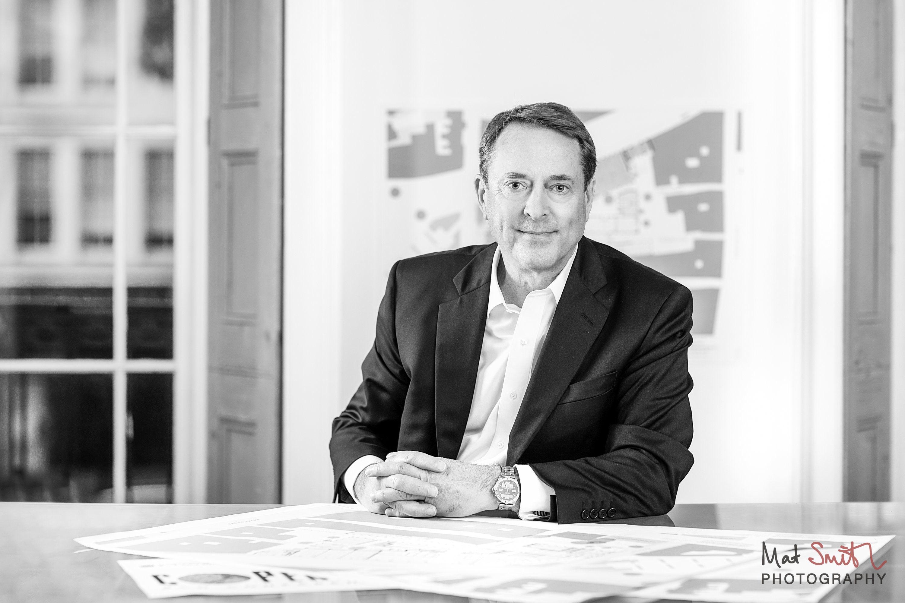 Black and white portrait of a man seated at a desk with plans in a bright office with tall windows, portrait by Mat Smith Photography, London