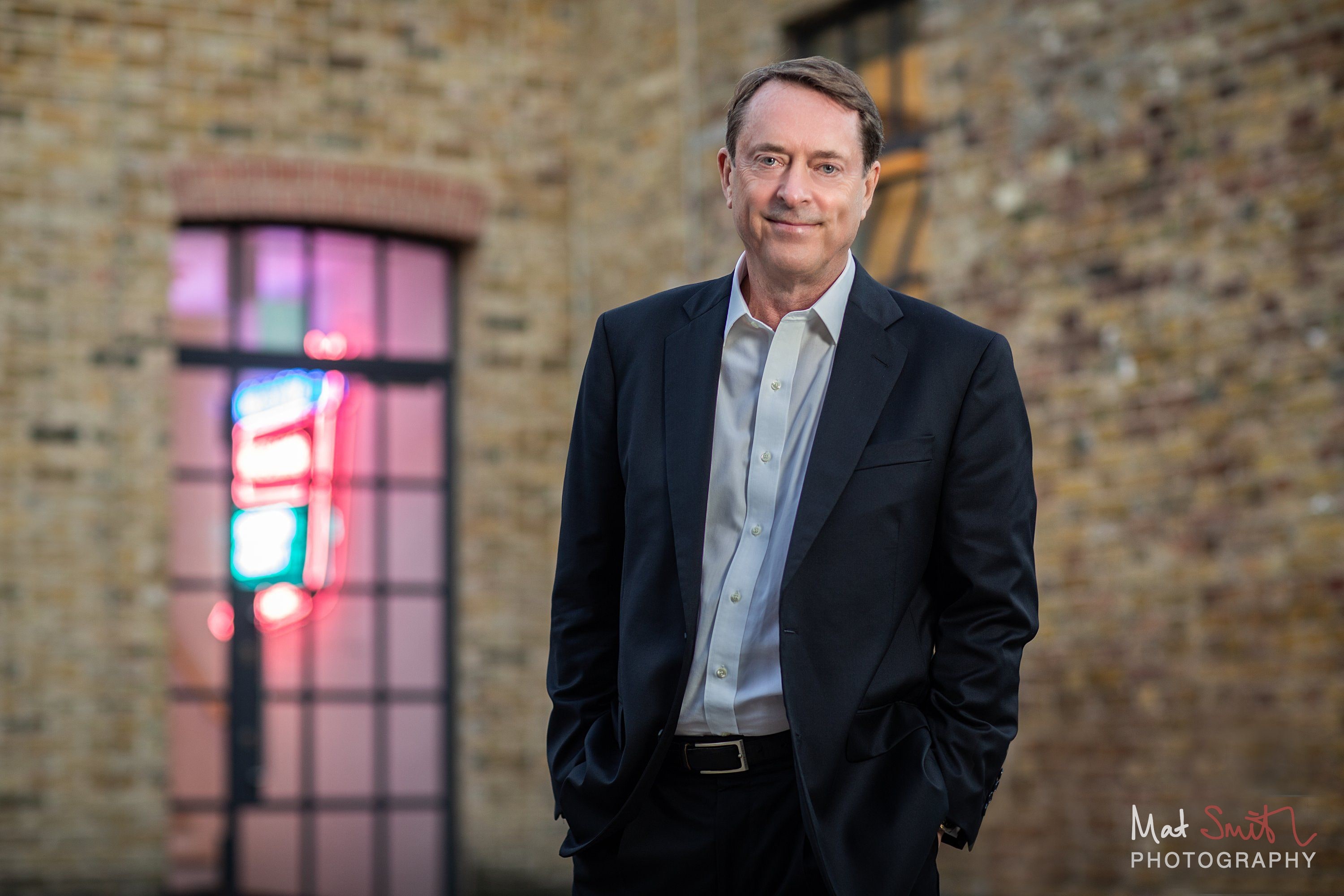 A man in a dark suit with hands in his pockets stands by a brick wall beside an arched window glowing with a neon sign, portrait by Mat Smith Photography, London