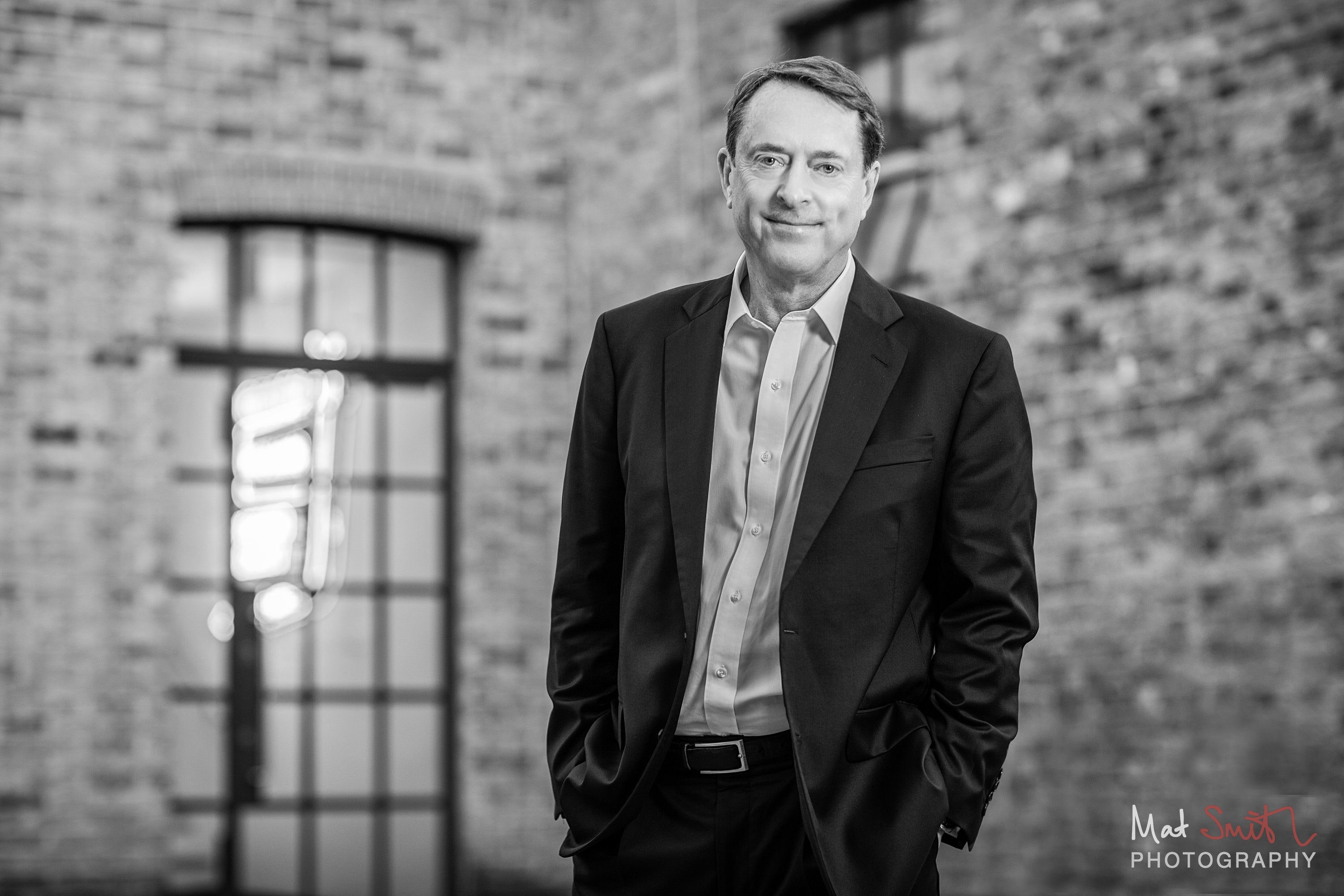 Black and white image of a man in a suit with hands in pockets beside a brick wall and arched window, portrait by Mat Smith Photography, London