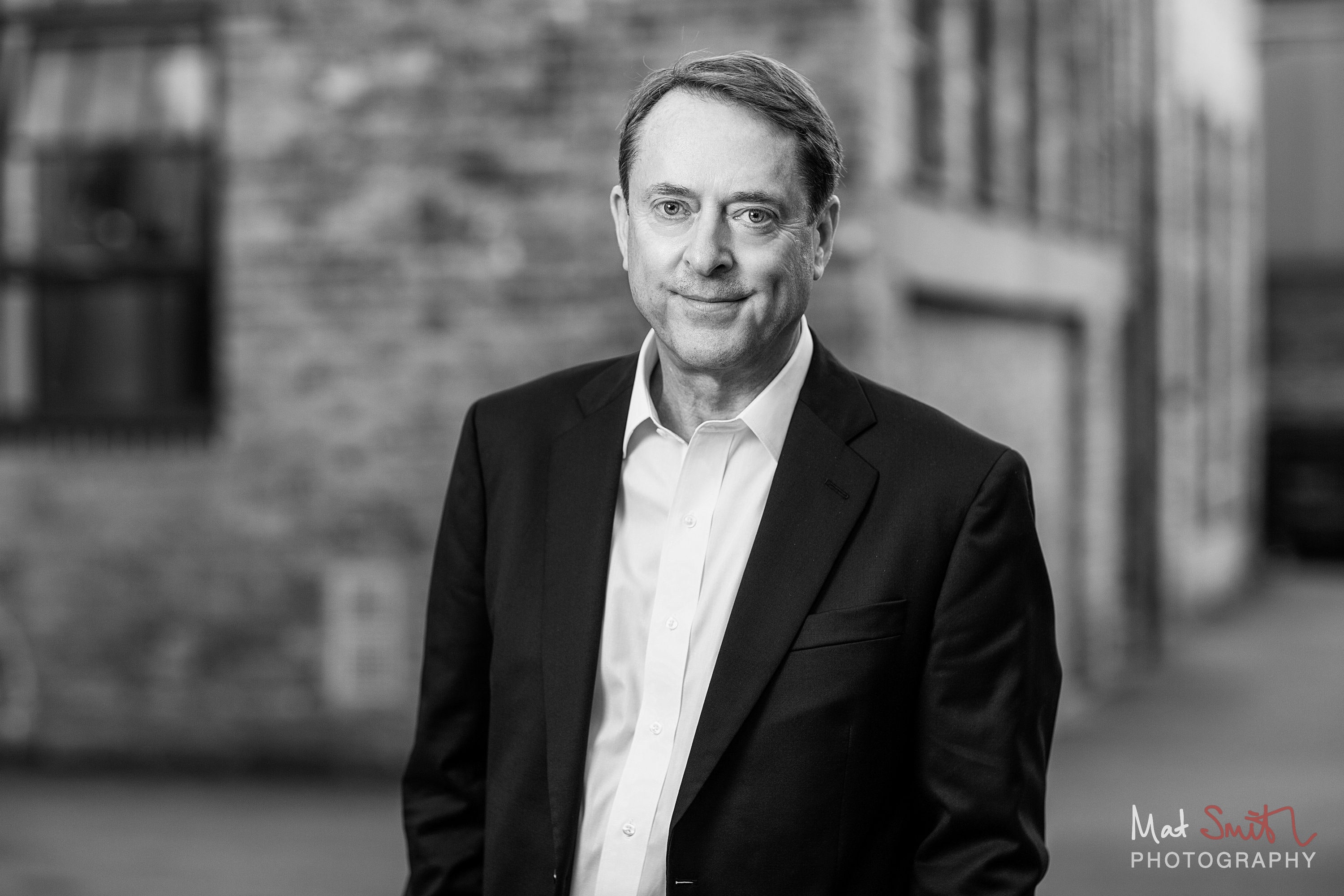 Black and white portrait of a smiling man in a suit in a lane between brick buildings, portrait by Mat Smith Photography, London