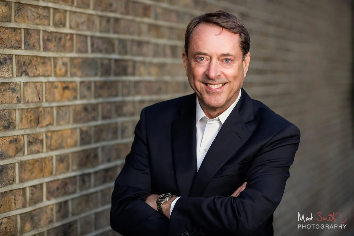 A smiling man in a dark suit with arms folded leans against a yellow brick wall, portrait by Mat Smith Photography, London