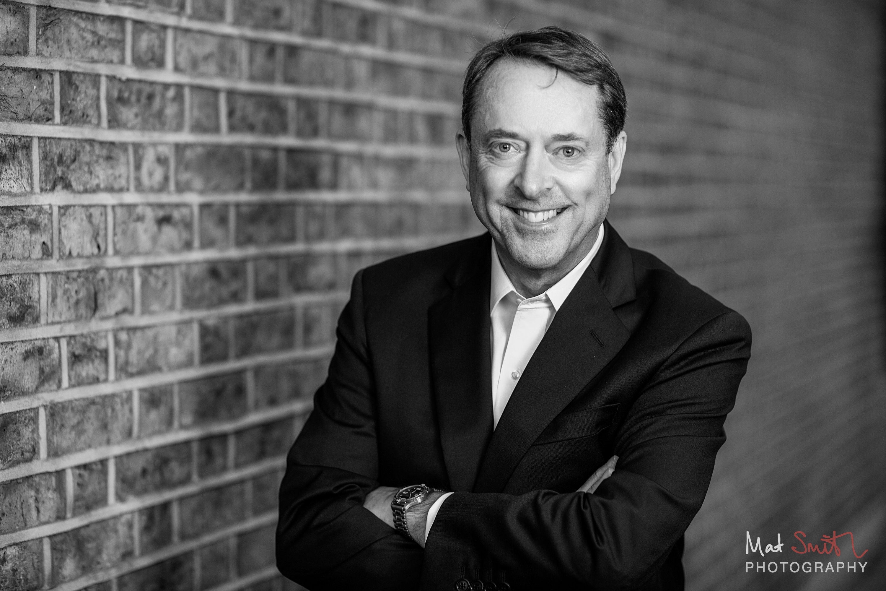 Black and white portrait of a smiling man in a suit with arms folded against a brick wall, portrait by Mat Smith Photography, London