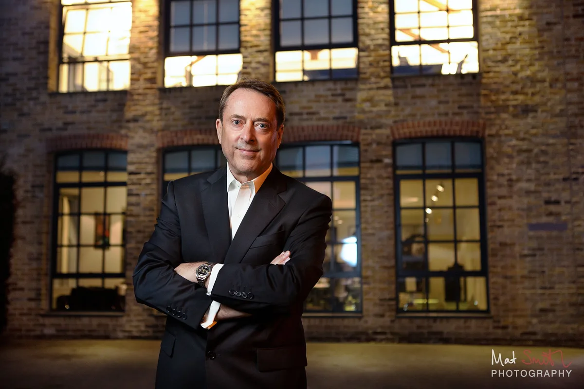 At dusk, a man in a dark suit stands with arms folded in front of a brick warehouse with lit windows, portrait by Mat Smith Photography, London
