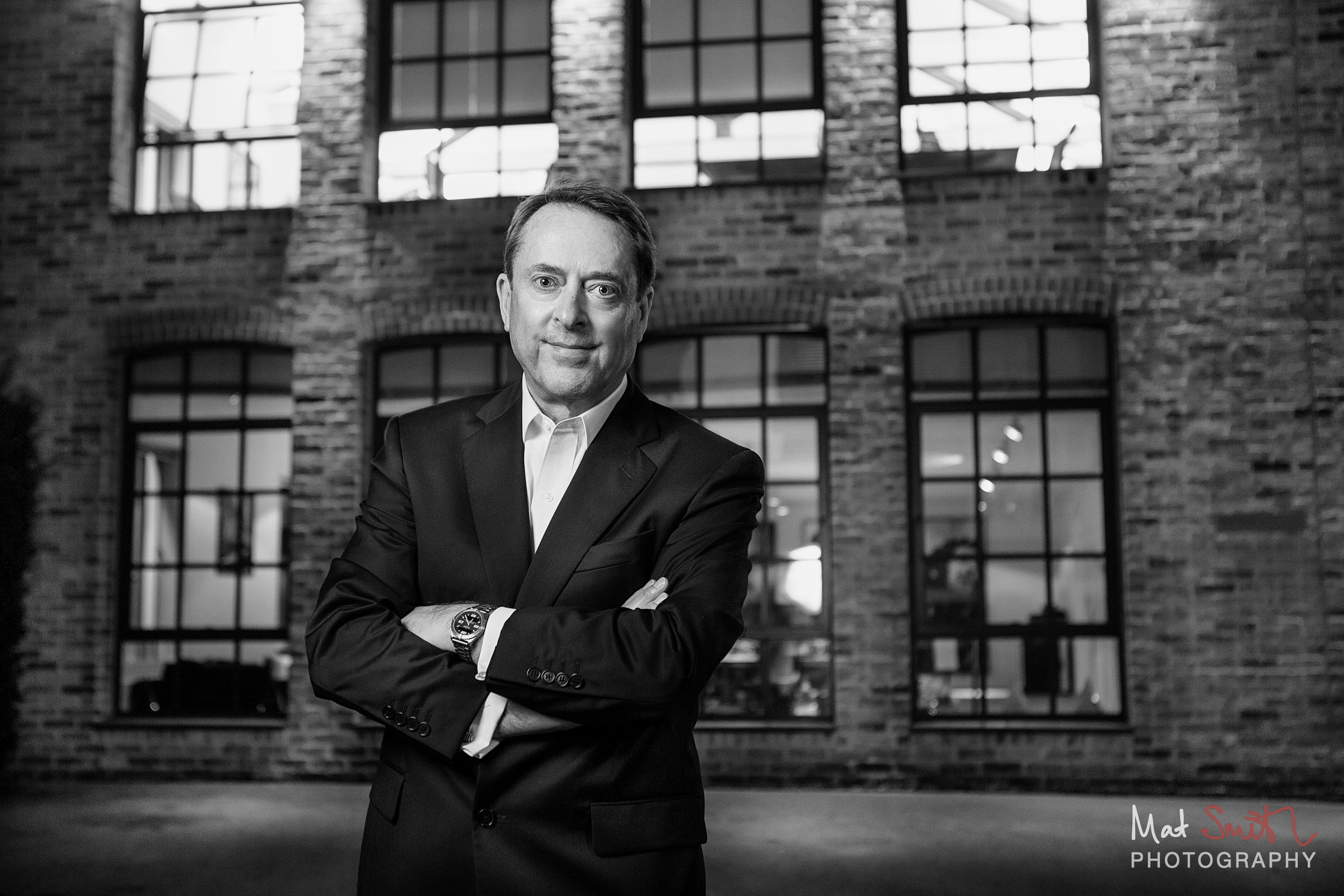 Black and white evening portrait of a man with arms folded in front of a brick warehouse with glowing windows, portrait by Mat Smith Photography, London