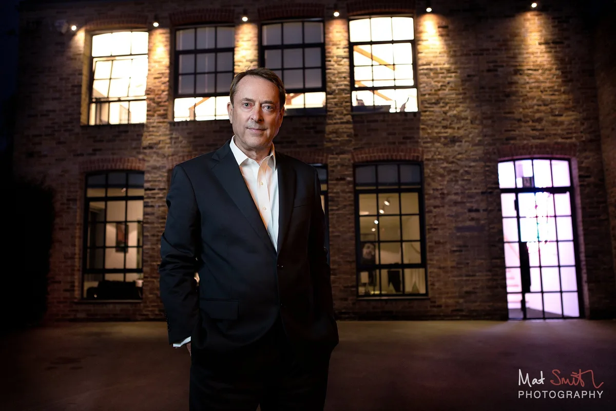 A man in a dark suit stands in a courtyard at dusk in front of a lit brick warehouse building with tall windows, portrait by Mat Smith Photography, London