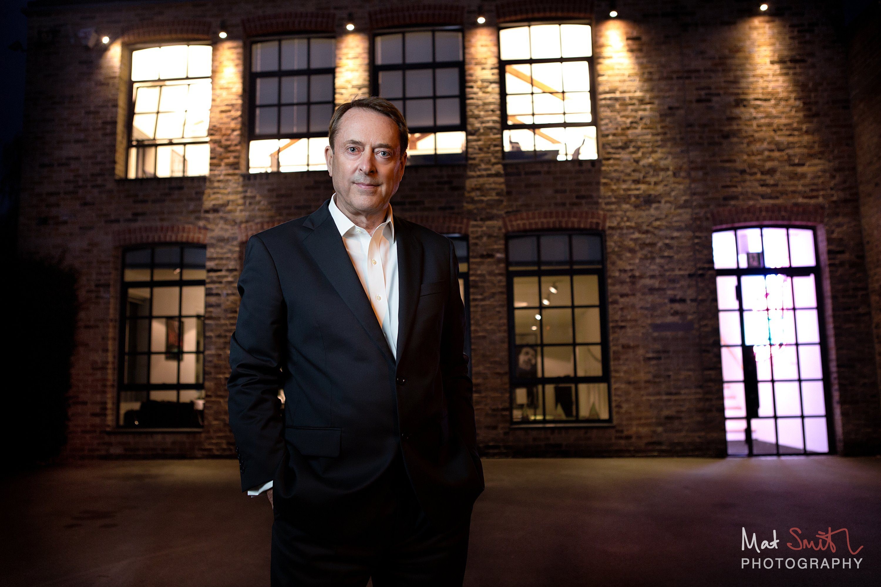 A man in a dark suit stands in a courtyard at dusk in front of a lit brick warehouse building with tall windows, portrait by Mat Smith Photography, London