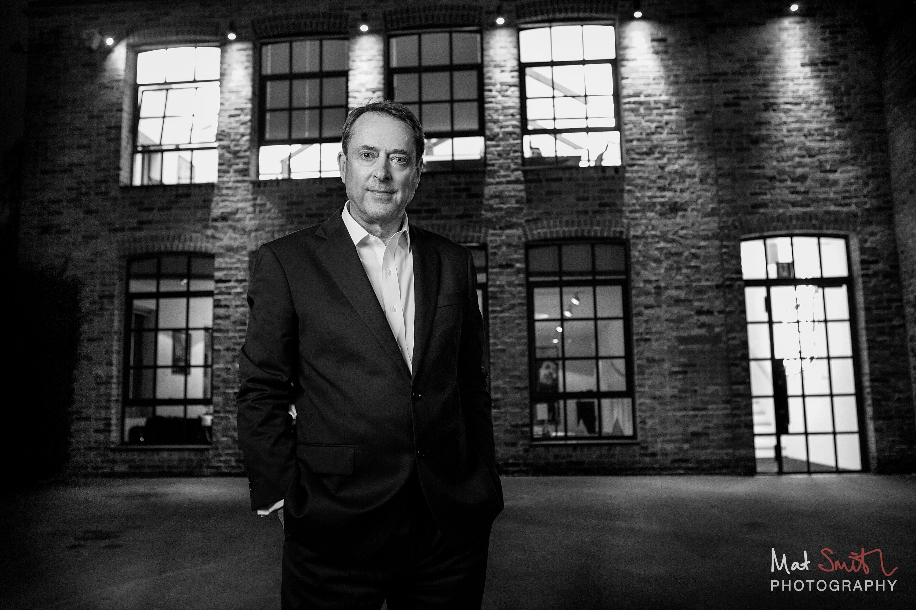Black and white image of a man in a suit standing in a courtyard at dusk before a lit warehouse facade, portrait by Mat Smith Photography, London