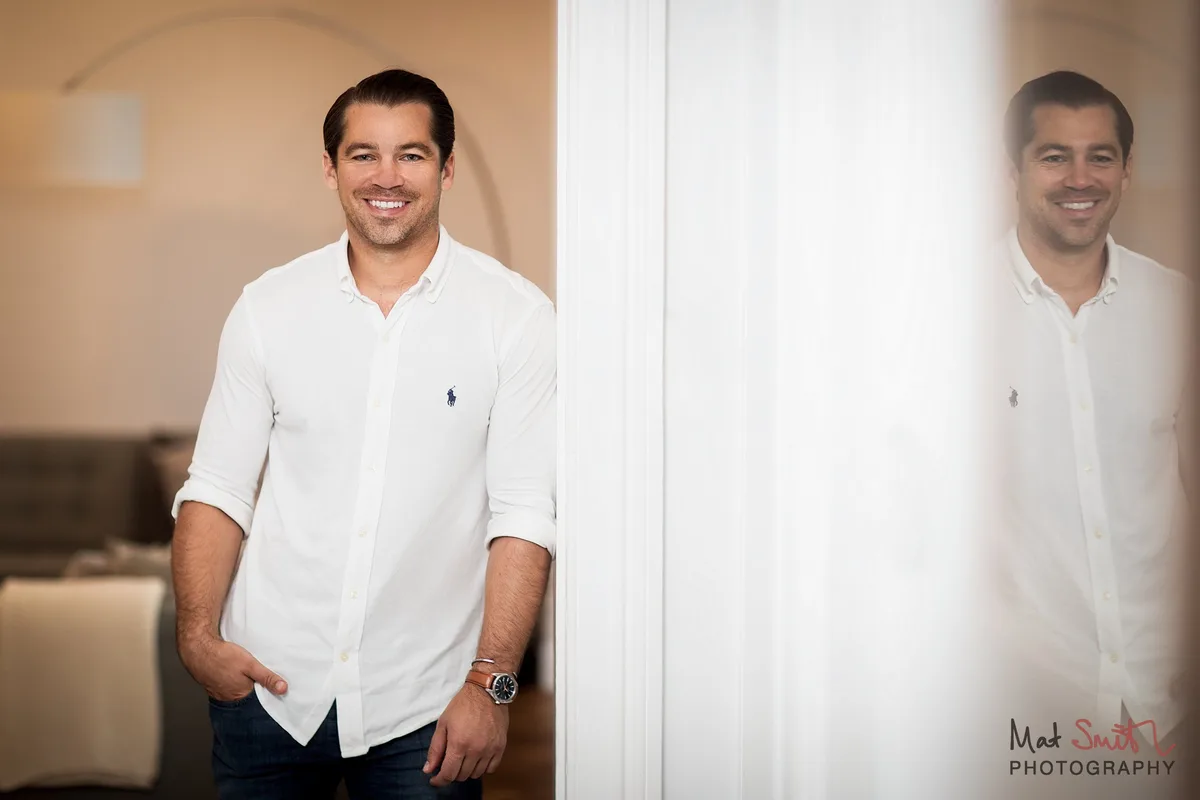 A smiling man with dark swept back hair in a white shirt and jeans, leaning in a doorway beside a frosted glass panel, portrait by Mat Smith Photography, London