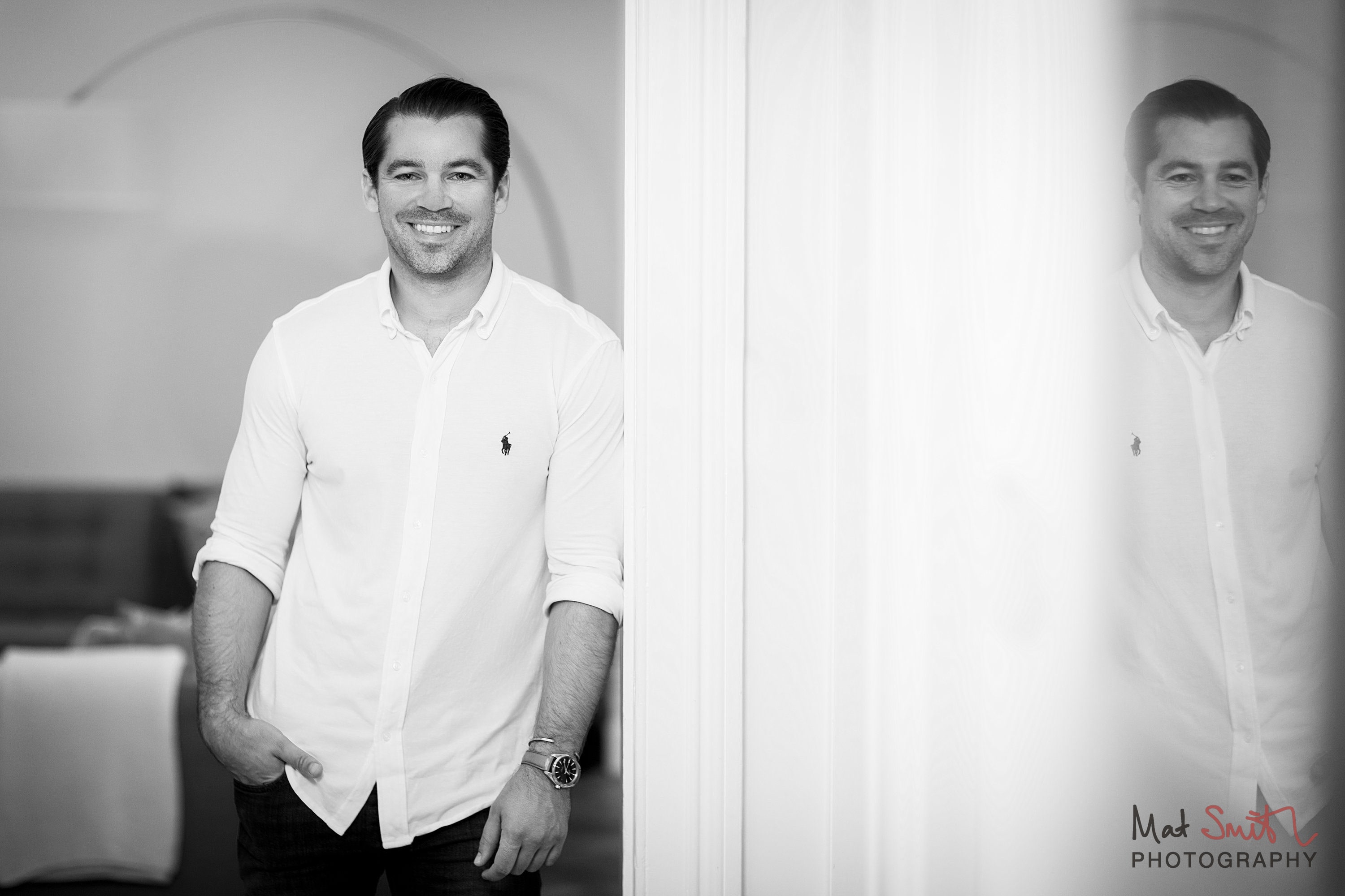 Black and white portrait of a smiling man in a white shirt leaning in a doorway beside a frosted glass panel, portrait by Mat Smith Photography, London