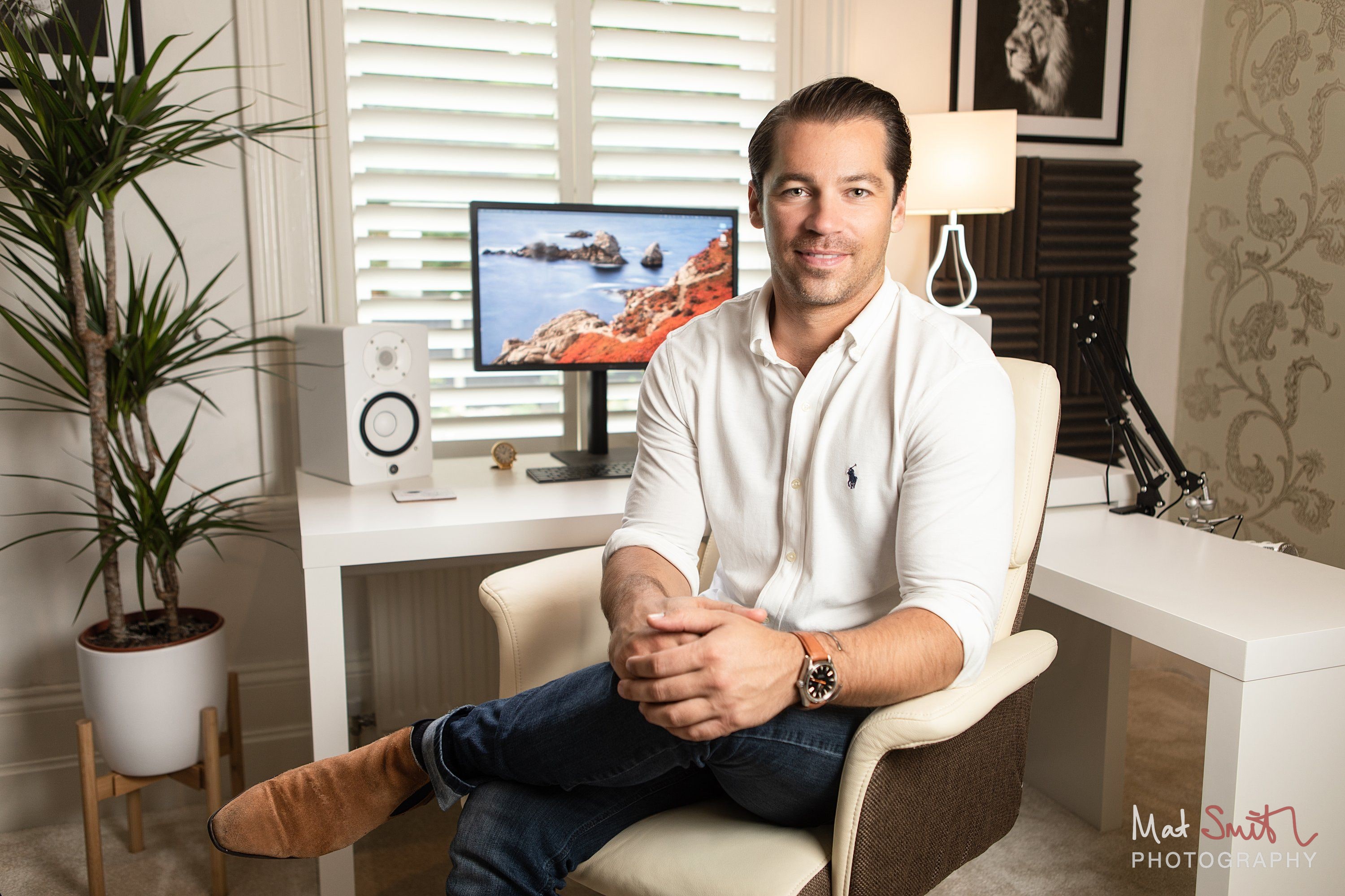 A man in a white shirt and jeans sitting cross legged in a cream chair at a white desk with a monitor, speaker and palm plant, portrait by Mat Smith Photography, London
