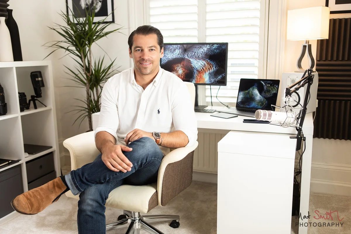 A man in a white shirt sitting in a cream office chair in a home studio with a monitor, laptop, microphone arm and shelving, portrait by Mat Smith Photography, London