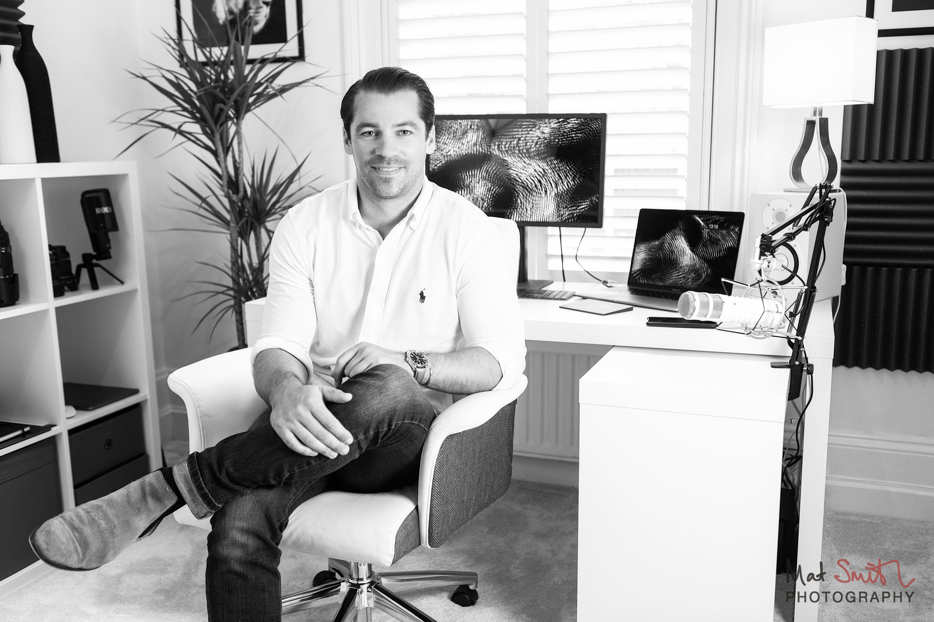 Black and white portrait of a man seated in a home office with a monitor, laptop and microphone arm, portrait by Mat Smith Photography, London