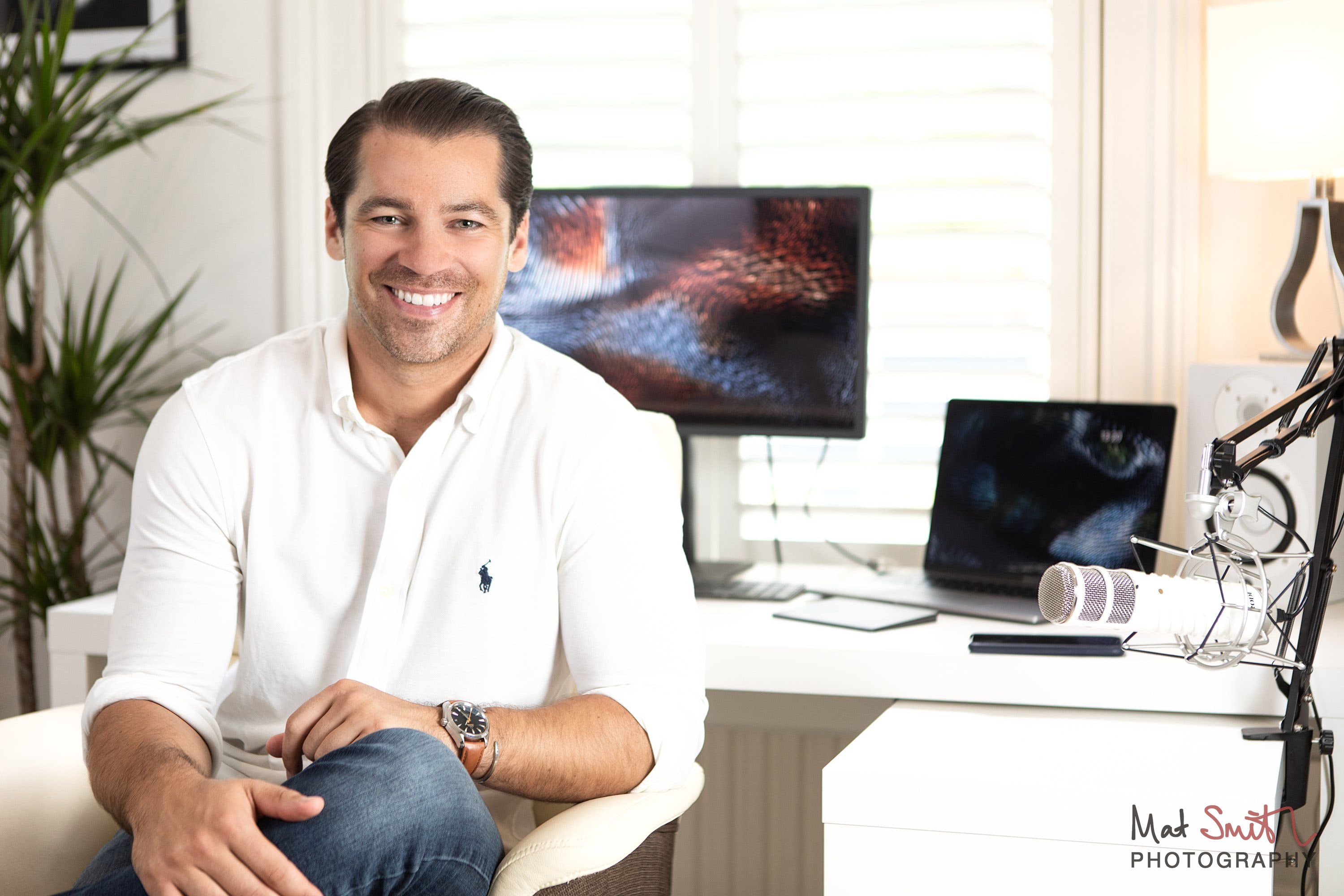 A smiling man in a white shirt seated at a desk, with a colourful monitor, laptop and microphone behind him, portrait by Mat Smith Photography, London