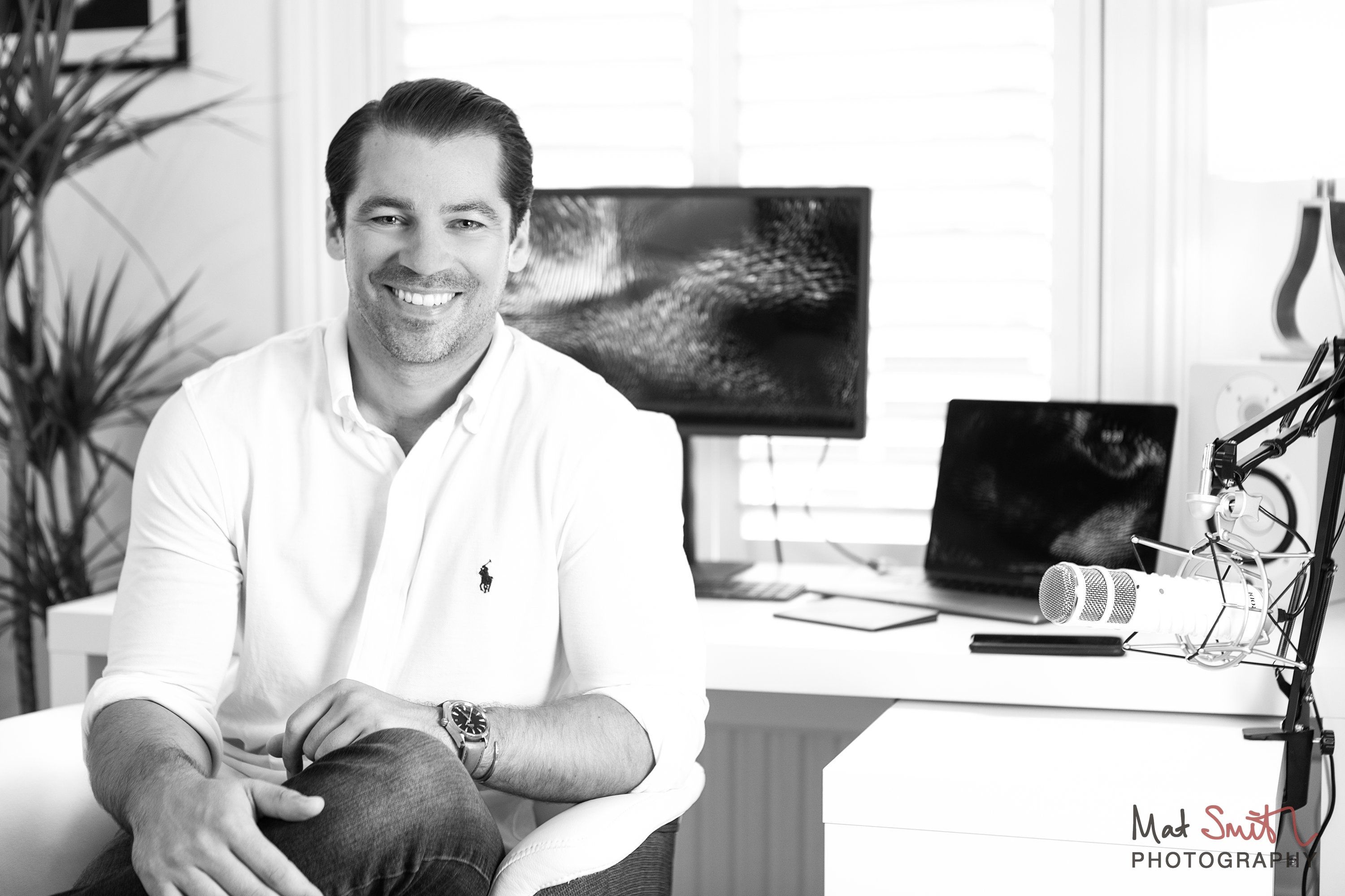 Black and white portrait of a smiling man in a white shirt seated in front of a desk with a monitor and laptop, portrait by Mat Smith Photography, London