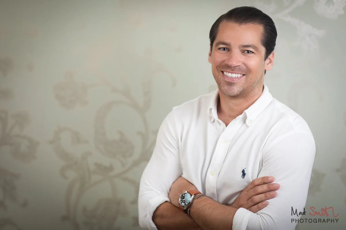 A smiling man in a white shirt with arms folded, framed to the right against pale patterned wallpaper, portrait by Mat Smith Photography, London