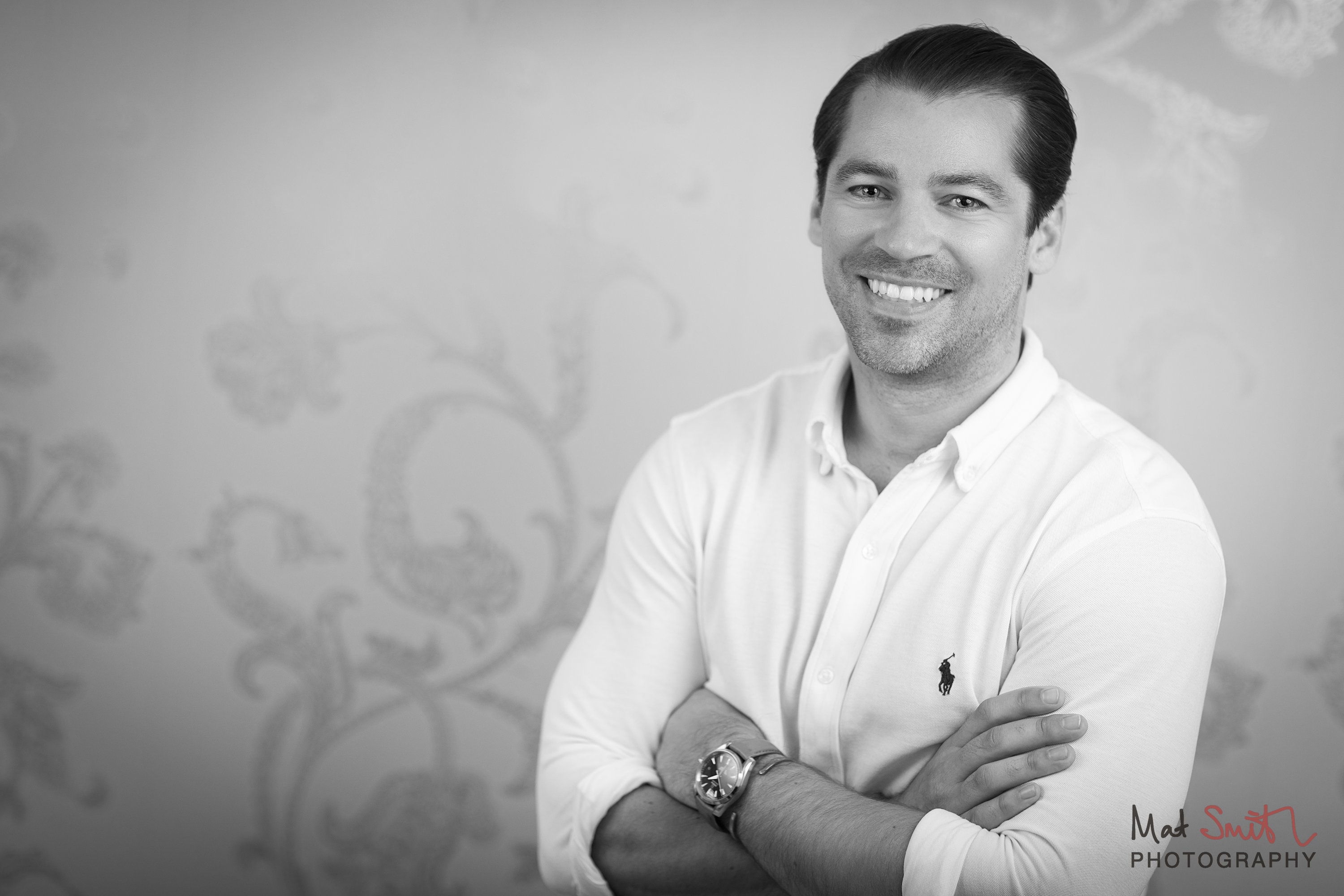 Black and white portrait of a smiling man with arms folded against patterned wallpaper, portrait by Mat Smith Photography, London