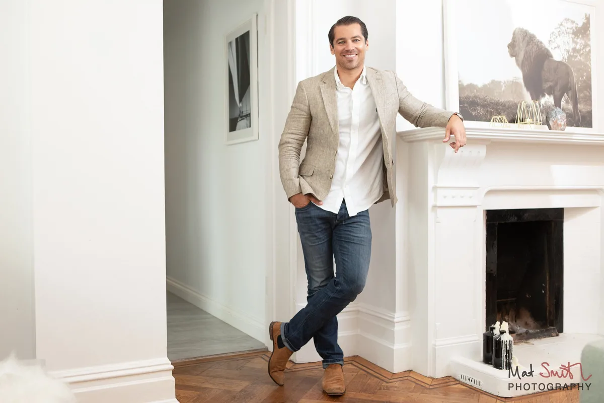 A man in a beige blazer and jeans leaning on a white fireplace mantel in a bright room, with a framed animal print above, portrait by Mat Smith Photography, London
