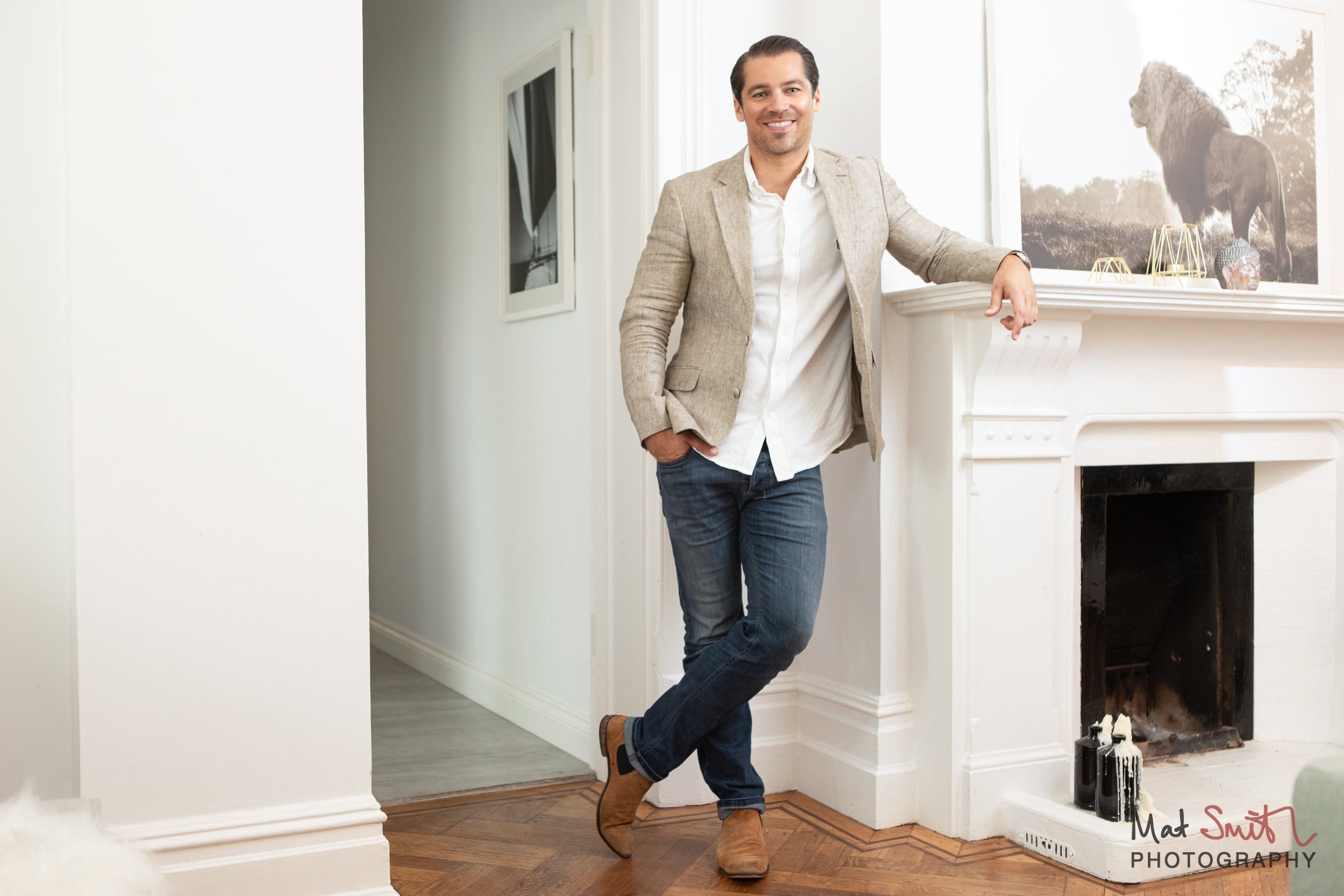 A man in a beige blazer and jeans leaning on a white fireplace mantel in a bright room, with a framed animal print above, portrait by Mat Smith Photography, London