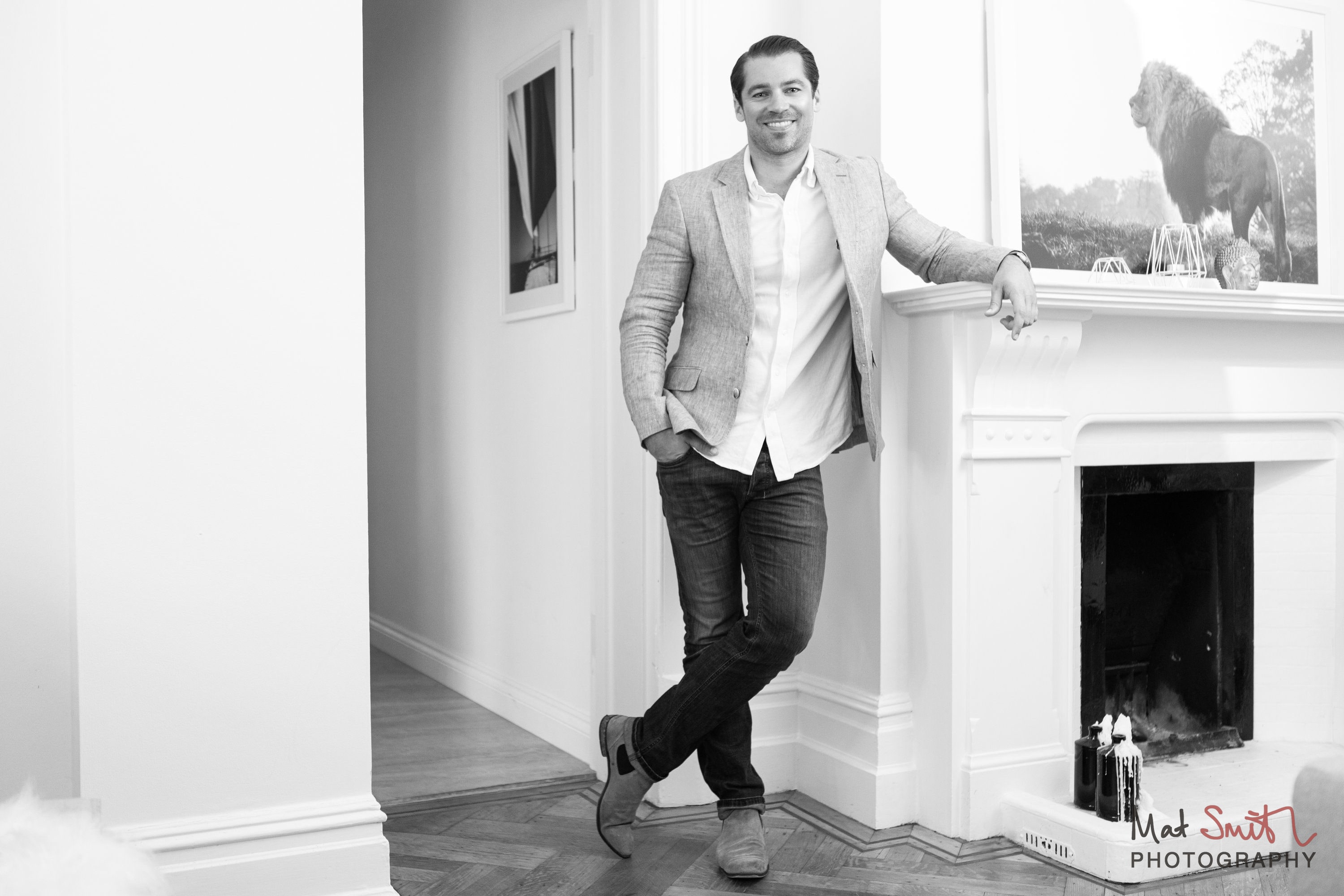 Black and white portrait of a man in a blazer leaning on a white fireplace mantel, legs crossed, portrait by Mat Smith Photography, London