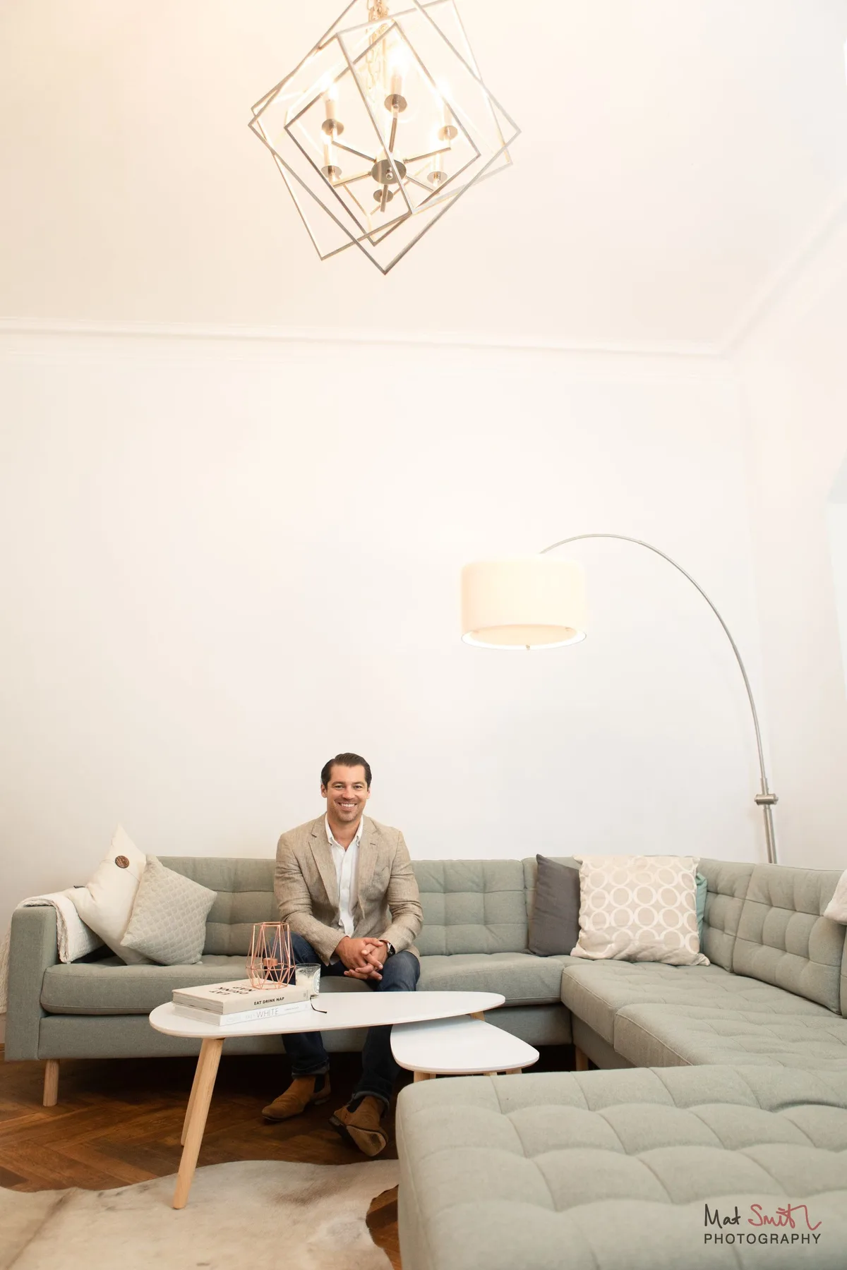 Vertical wide portrait of a man in a blazer sitting on a large grey corner sofa beneath a geometric chandelier and arc lamp, portrait by Mat Smith Photography, London