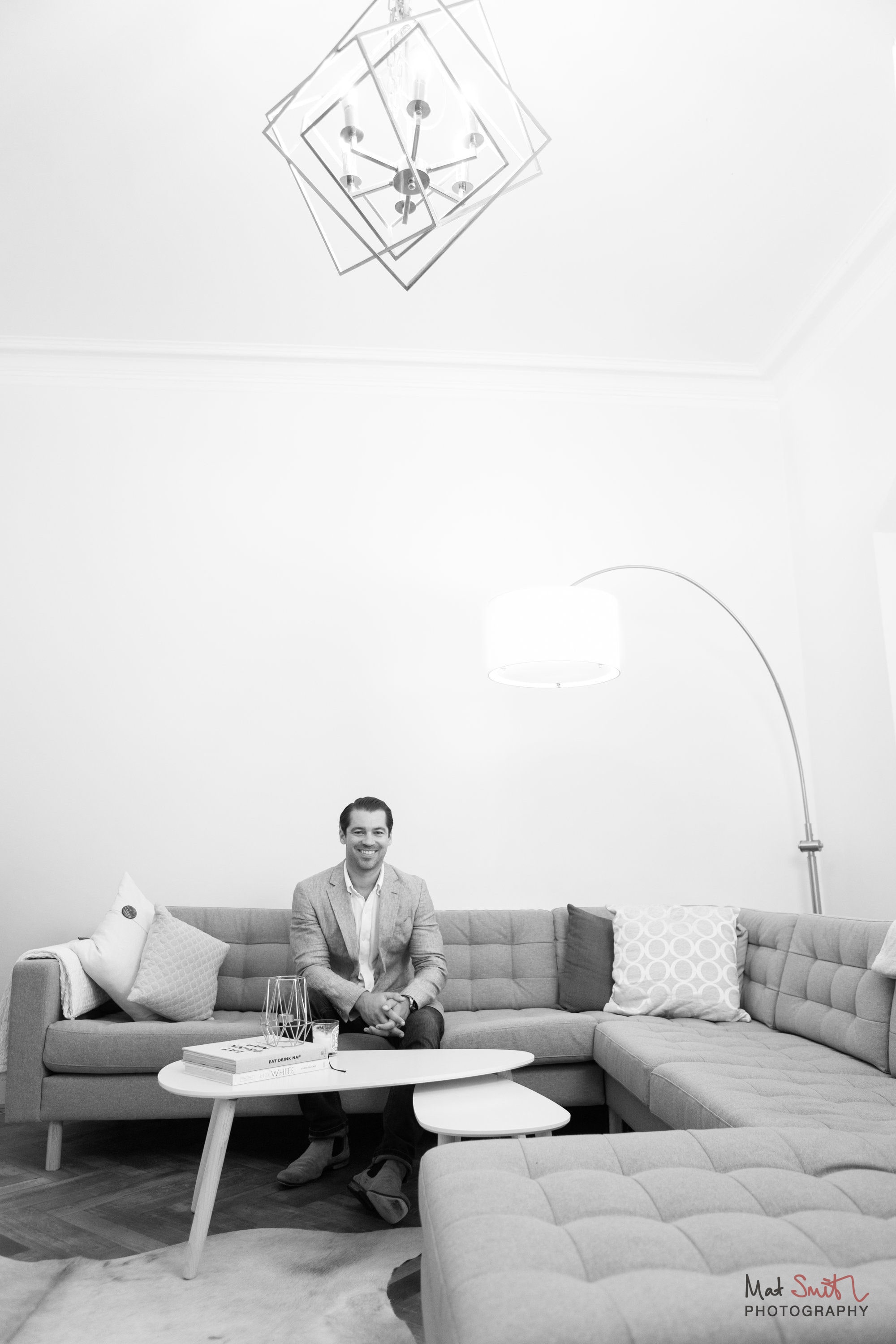 Black and white vertical portrait of a man on a large corner sofa under a geometric chandelier, portrait by Mat Smith Photography, London