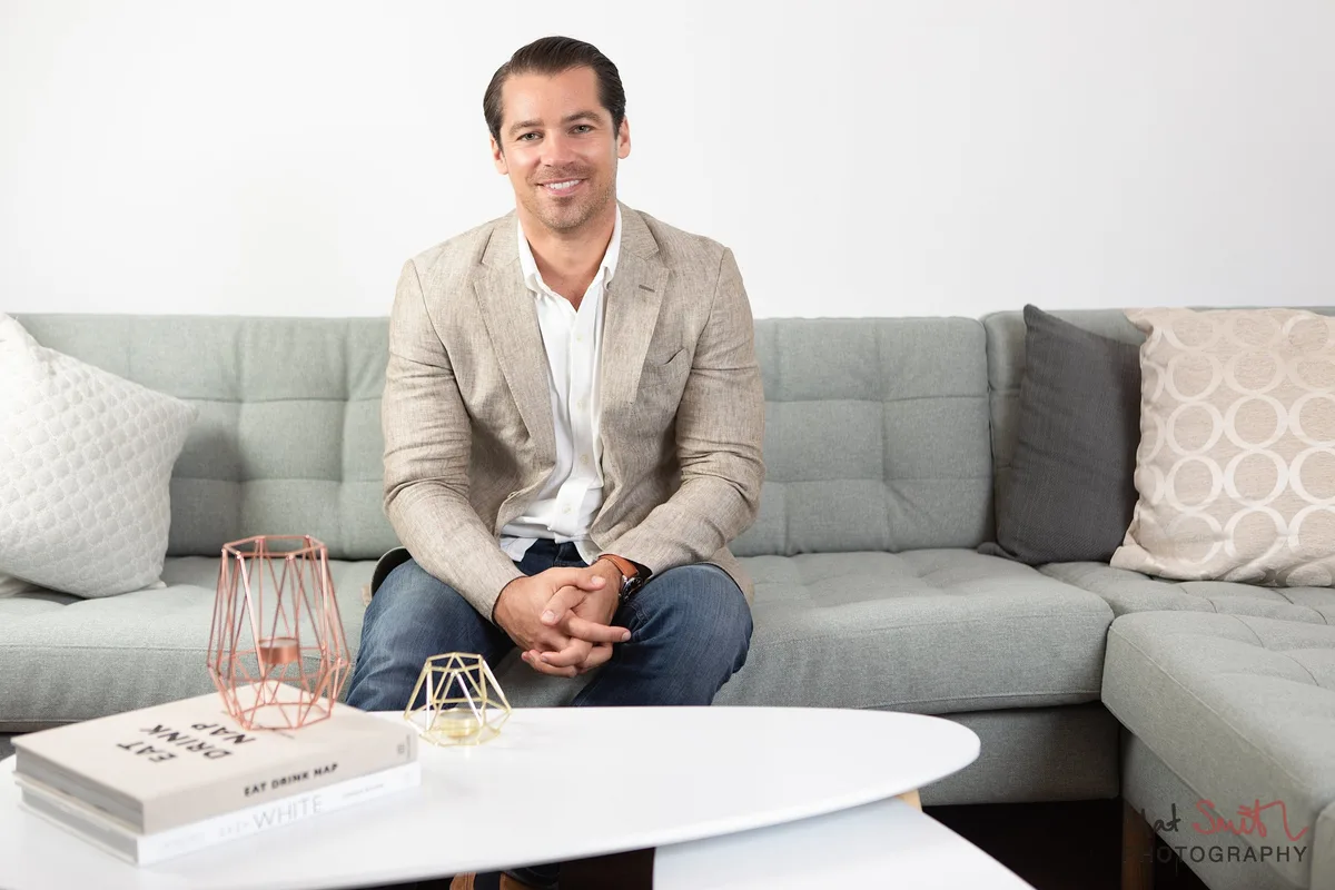 A smiling man in a beige blazer sitting on a grey sofa behind a white coffee table with books and candle holders, portrait by Mat Smith Photography, London