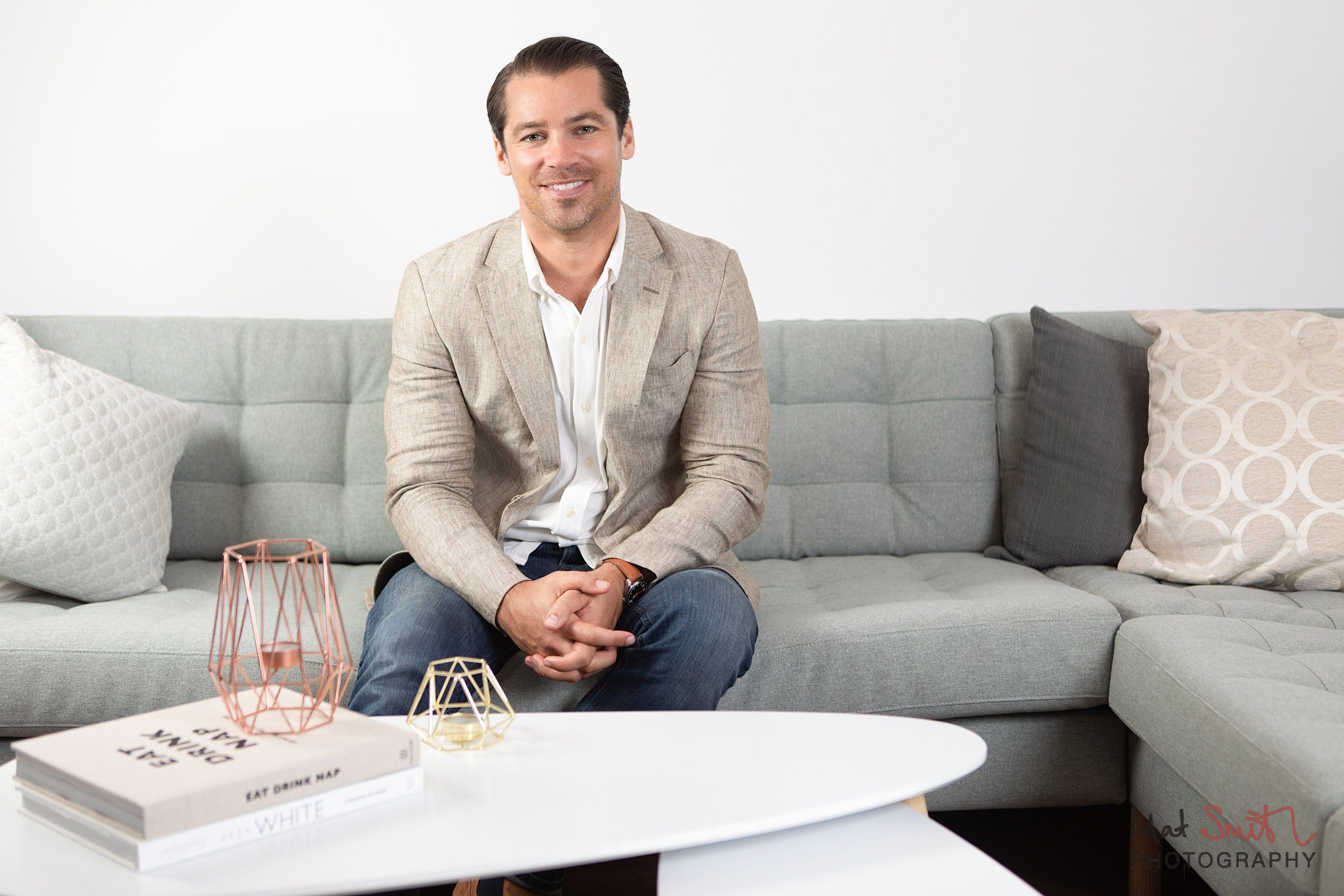 A smiling man in a beige blazer sitting on a grey sofa behind a white coffee table with books and candle holders, portrait by Mat Smith Photography, London