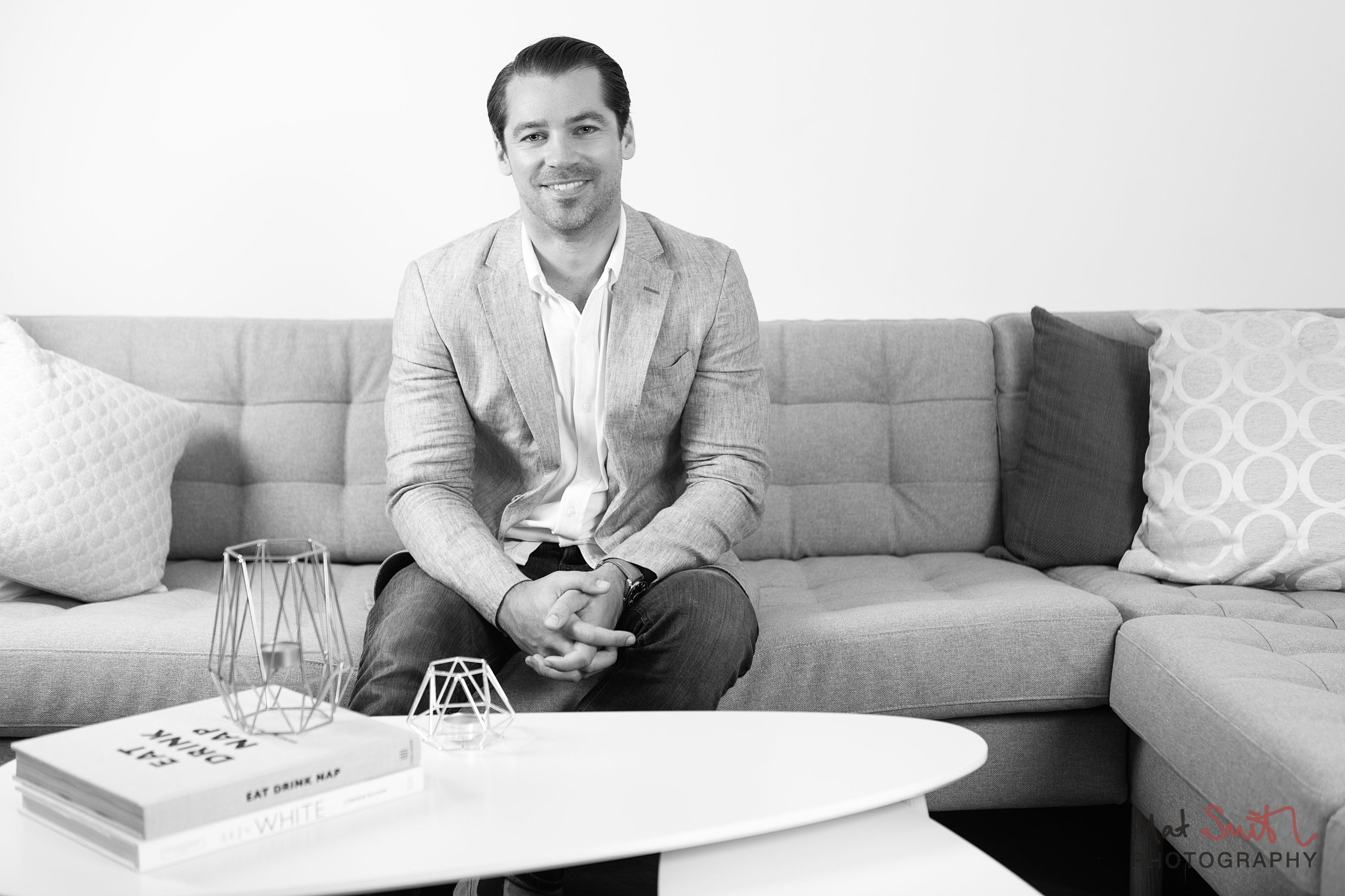 Black and white portrait of a man on a sofa behind a coffee table with books and geometric candle holders, portrait by Mat Smith Photography, London