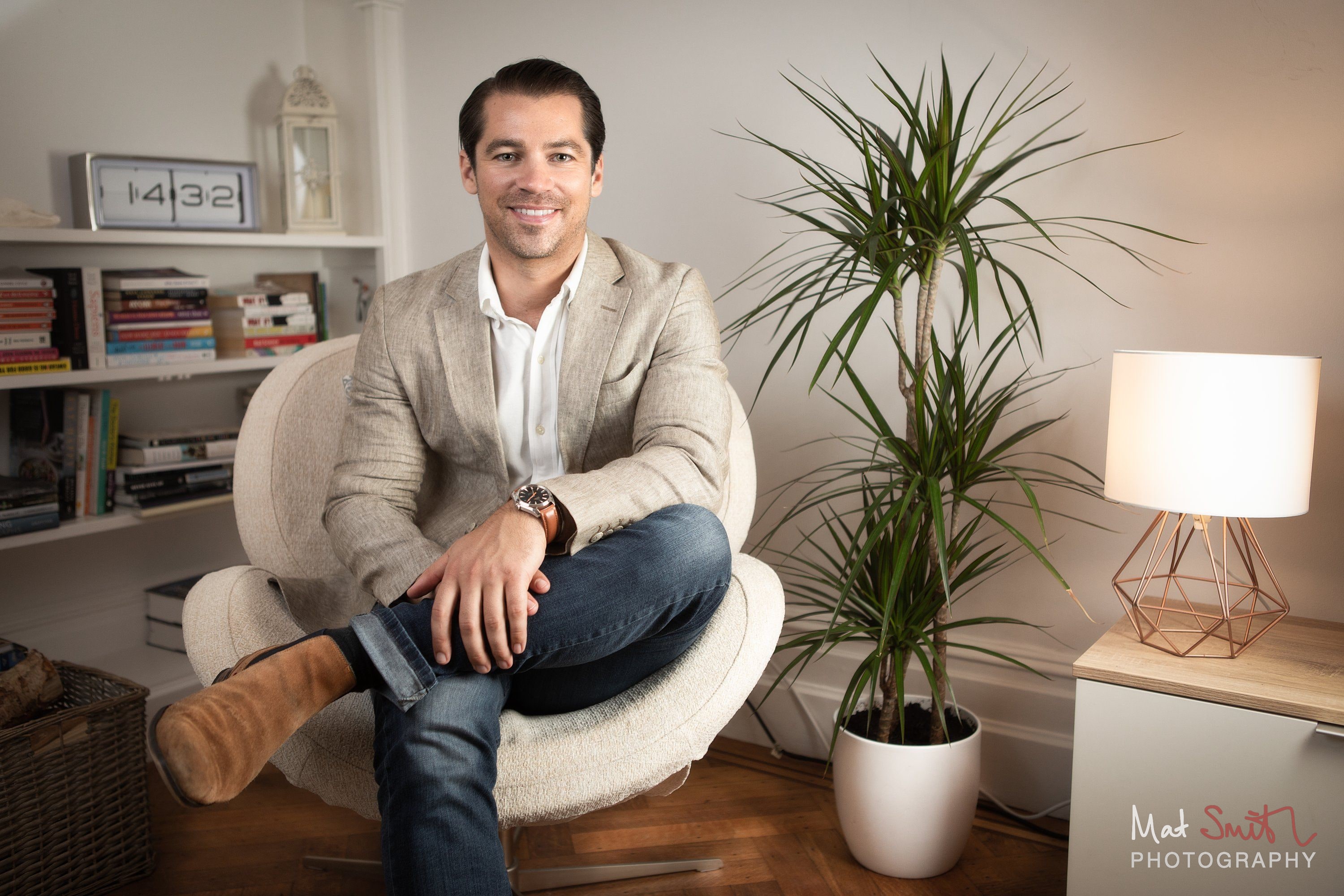 A man in a beige blazer and jeans sitting cross legged in a white armchair beside a tall plant, bookshelves and a lamp, portrait by Mat Smith Photography, London